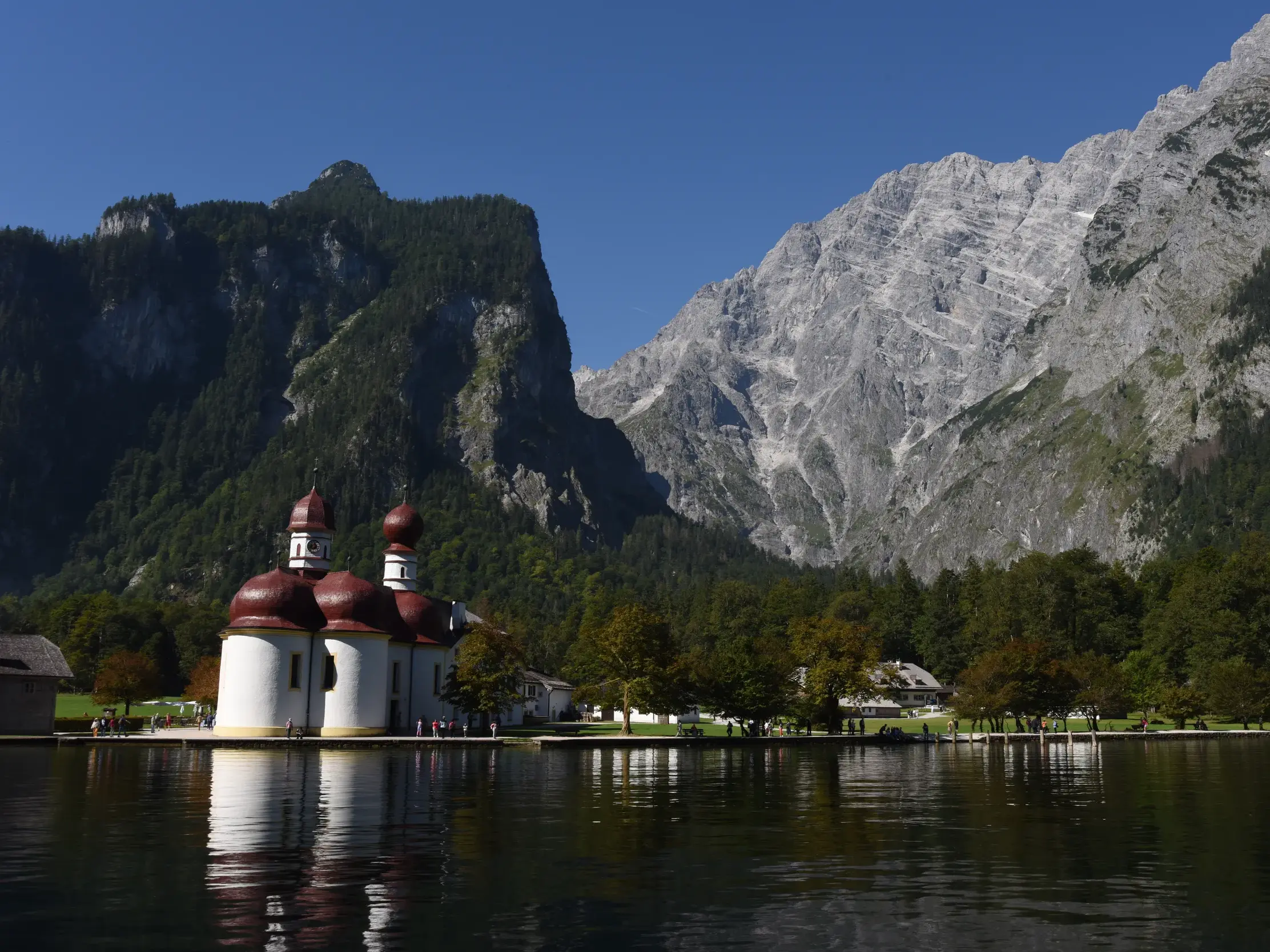 Die Wallfahrtskirche St. Bartholomä am Königssee spiegelt sich im Wasser vor den massiven Felswänden der Berchtesgadener Alpen.