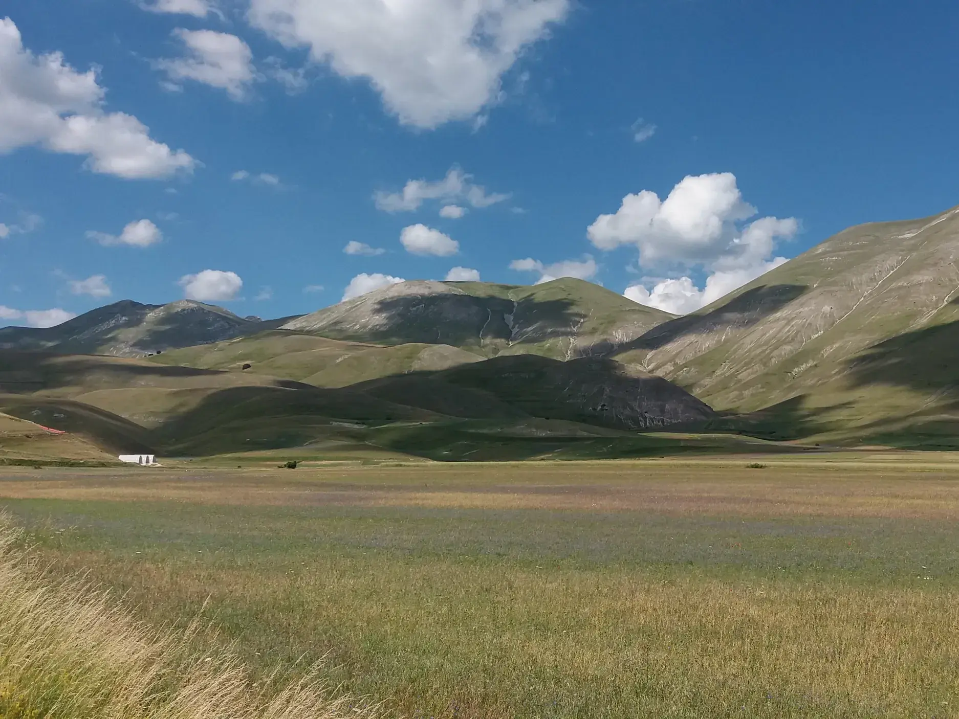 Weite Wiesen der Hochebene Piano Grande mit den grünen Gipfeln des Nationalparks Monti Sibillini unter blauem Himmel.