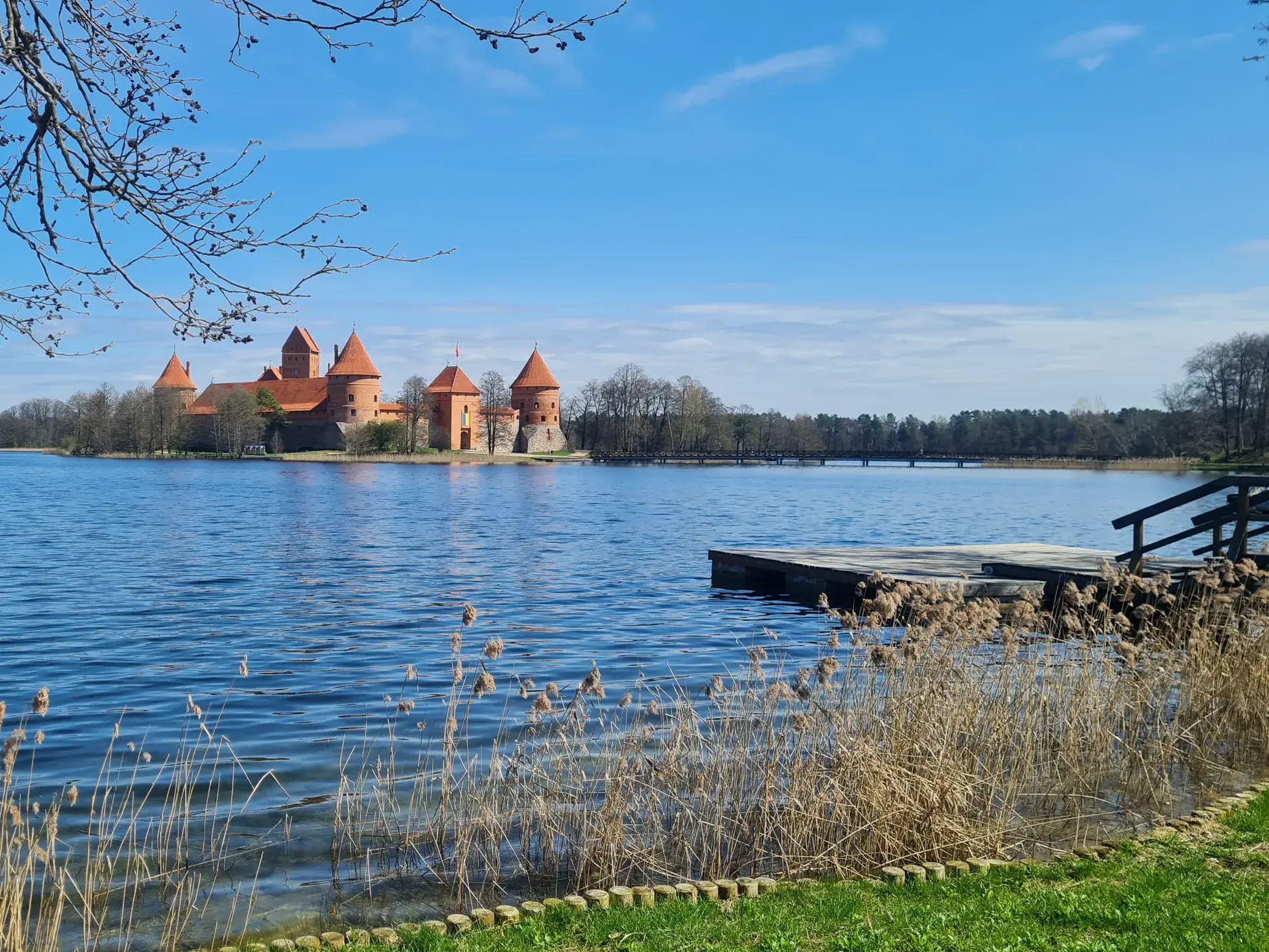Blick über den blauen Galve-See auf das Wasserschloss Trakai in Litauen, im Vordergrund ein Holzsteg am schilfbewachsenen Ufer.