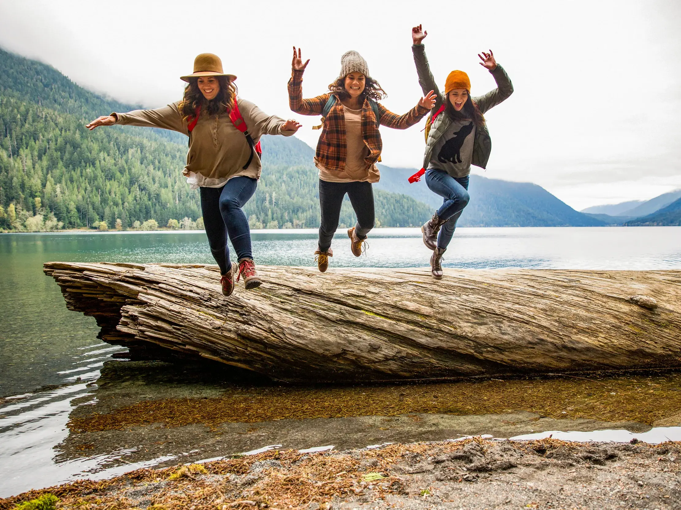 Drei junge Frauen in Wanderkleidung springen voller Freude von einem Baumstamm an einem Bergsee in den kanadischen Rockies.