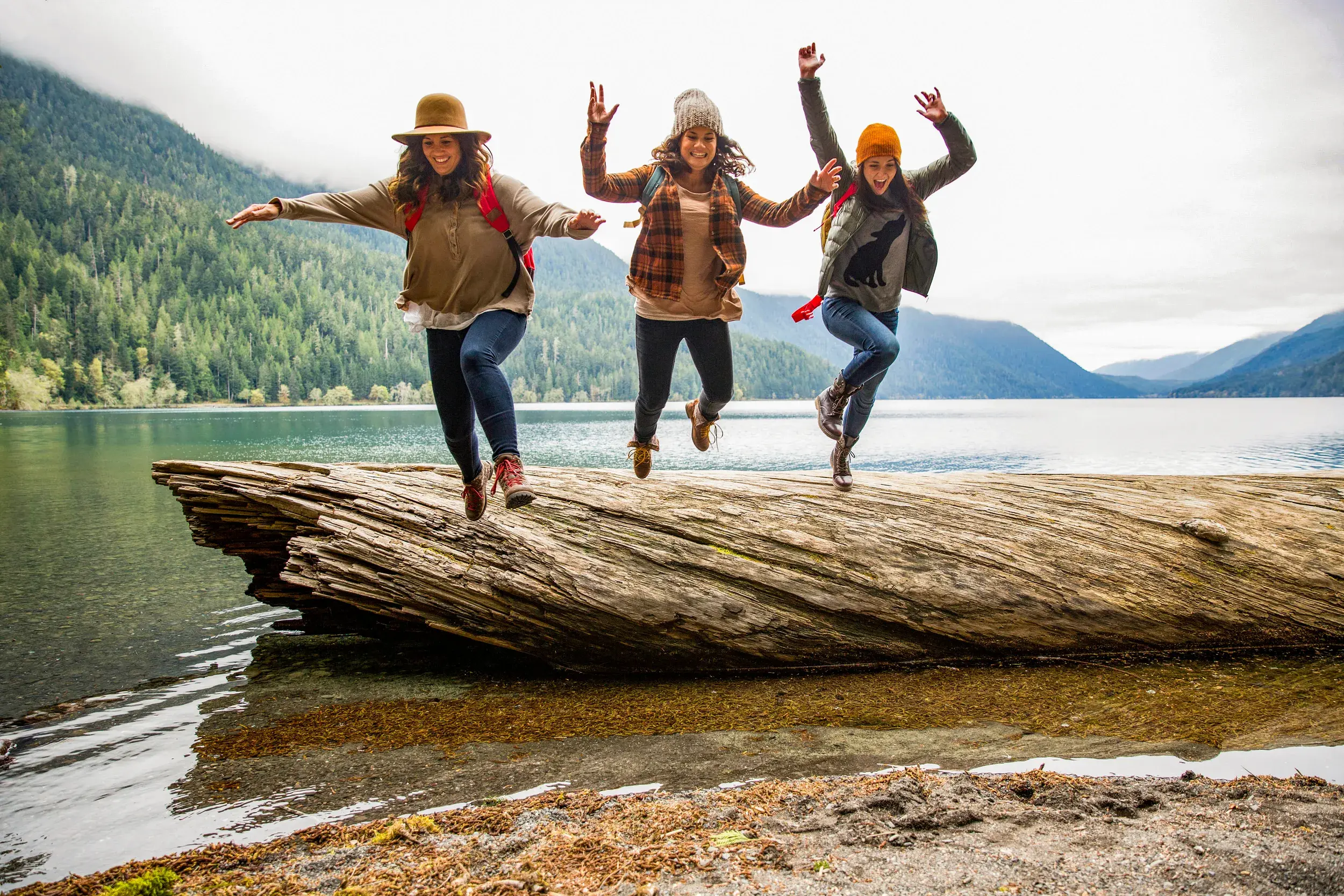 Drei junge Frauen in Wanderkleidung springen voller Freude von einem Baumstamm an einem Bergsee in den kanadischen Rockies.