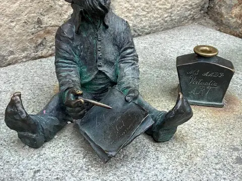 Nahaufnahme einer Bronzestatue eines der berühmten Breslauer Zwerge, die auf dem Steinboden sitzt und schreibt.