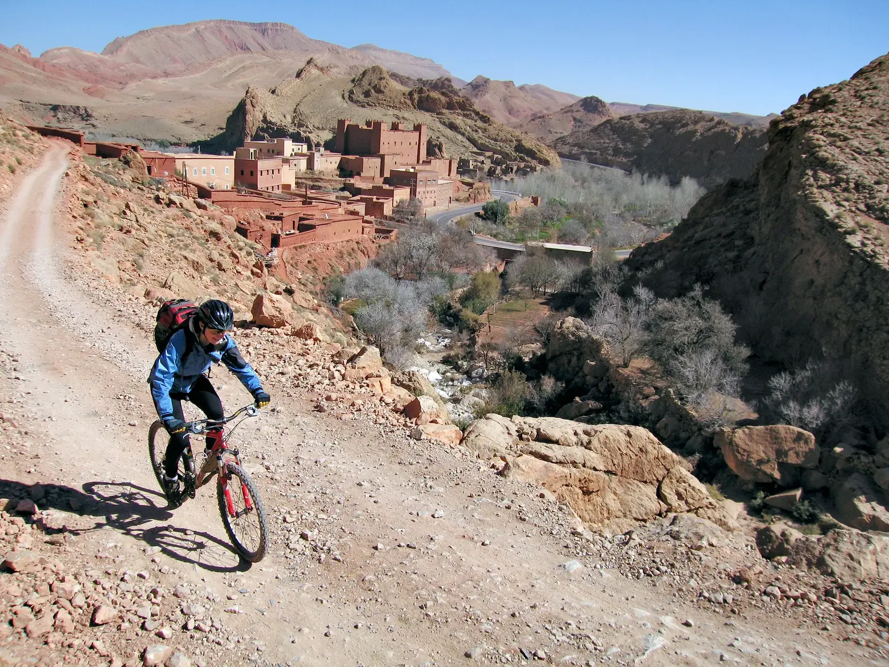 Mountainbiker auf einer Schotterpiste oberhalb der Dades-Schlucht mit Blick auf eine traditionelle Kasbah im Hohen Atlas.