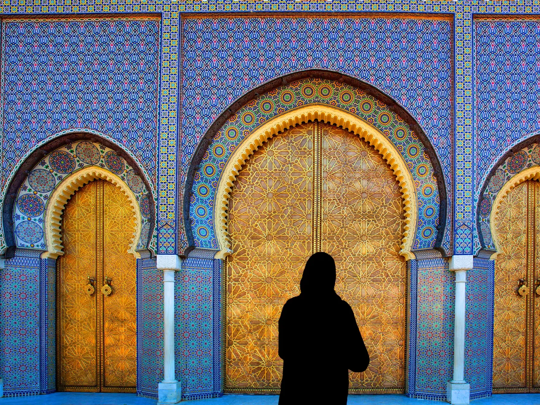 Silhouette einer Frau vor den prachtvollen, goldenen Toren des Königspalastes in Fes, umgeben von blauen Zellij-Mosaiken.