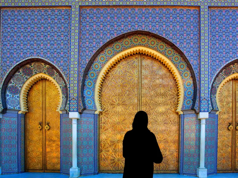 Silhouette einer Frau vor den prachtvollen, goldenen Toren des Königspalastes in Fes, umgeben von blauen Zellij-Mosaiken.
