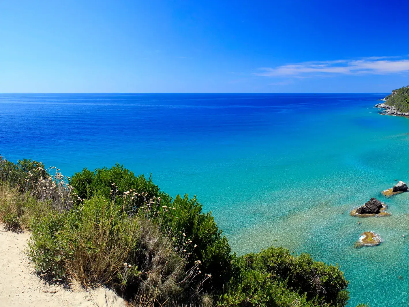 Panoramablick vom Korfu-Trail auf das türkisblaue Meer und die grüne, hügelige Küstenlinie Korfus an einem sonnigen Tag.