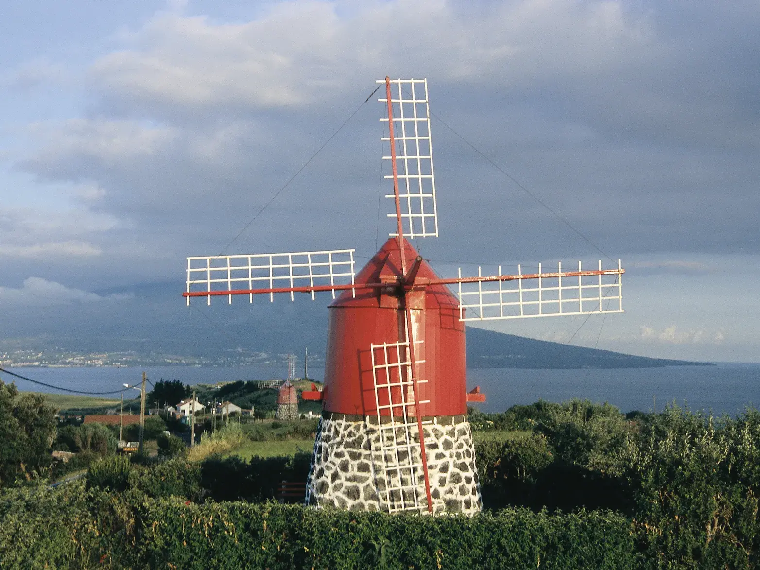 Traditionelle rote Windmühle auf der Azoreninsel Faial blickt über die grüne Küste zum Vulkan Pico am Horizont.
