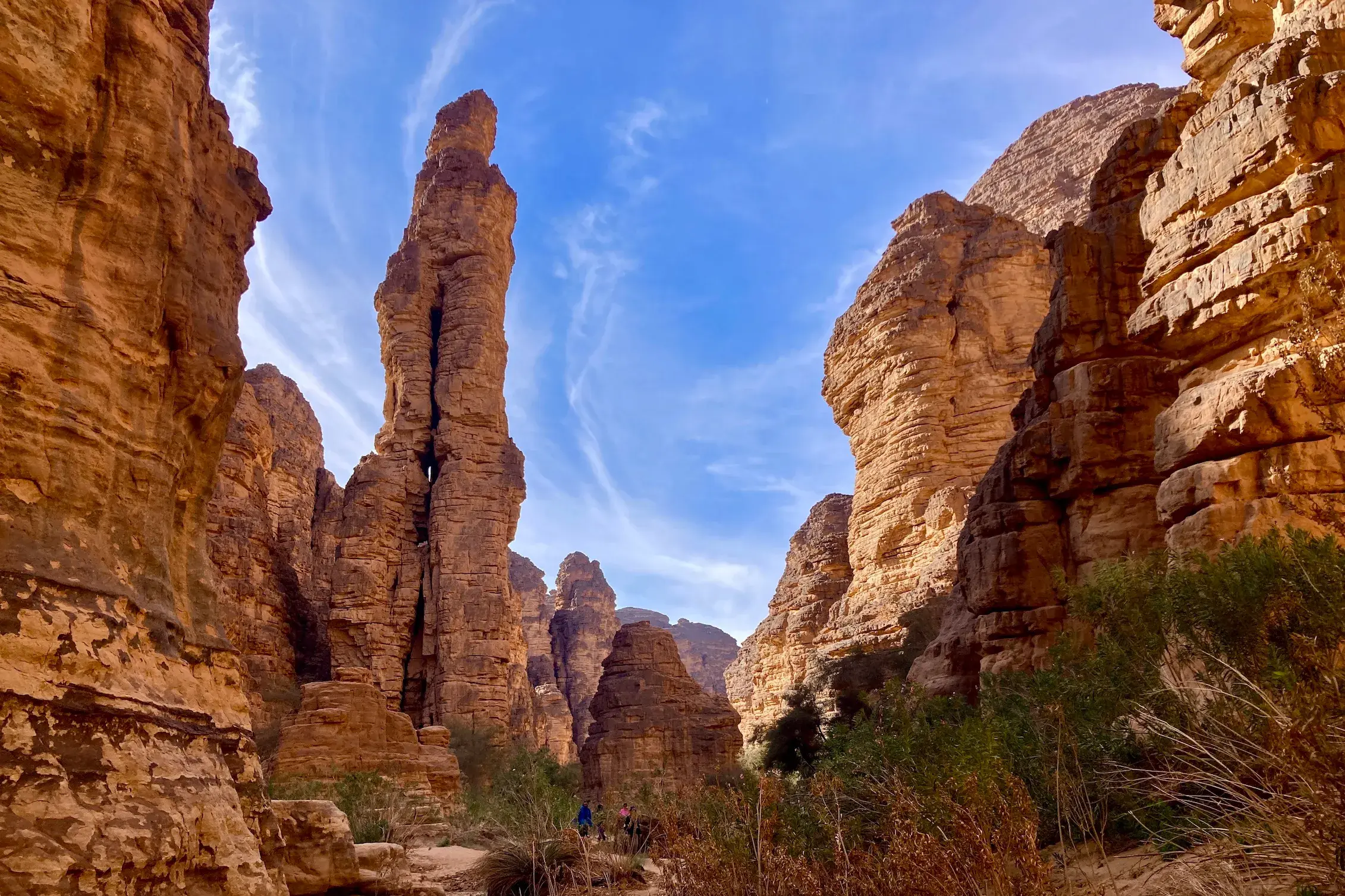 Eine Trekkinggruppe wandert durch eine Felsschlucht im Tassili n'Ajjer Nationalpark in Algerien, vorbei an einer hohen Felsnadel.