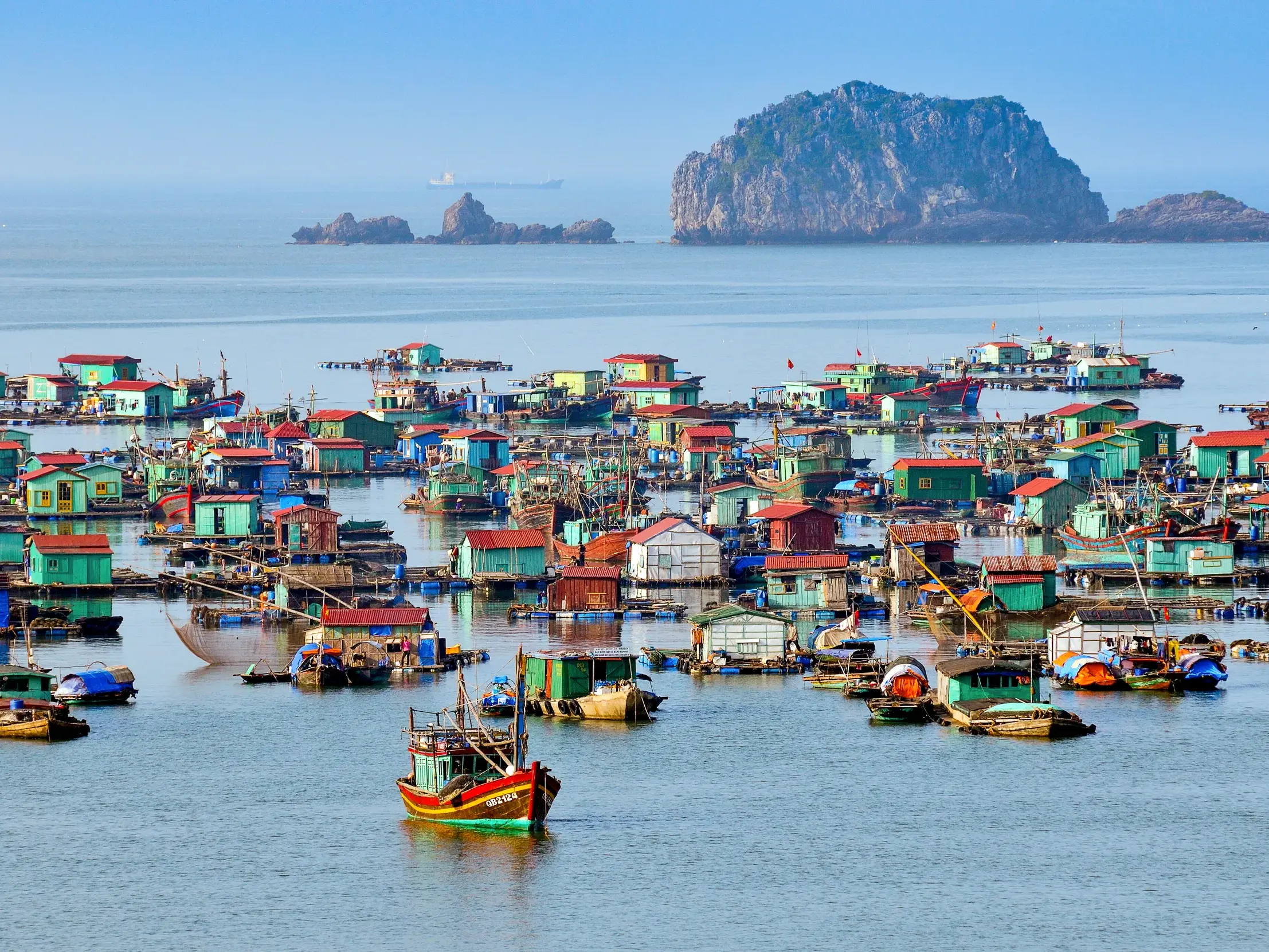Bunte, schwimmende Häuser und Fischerboote des Dorfes Cai Beo in der Halongbucht, Vietnam, vor einer großen Karstinsel.