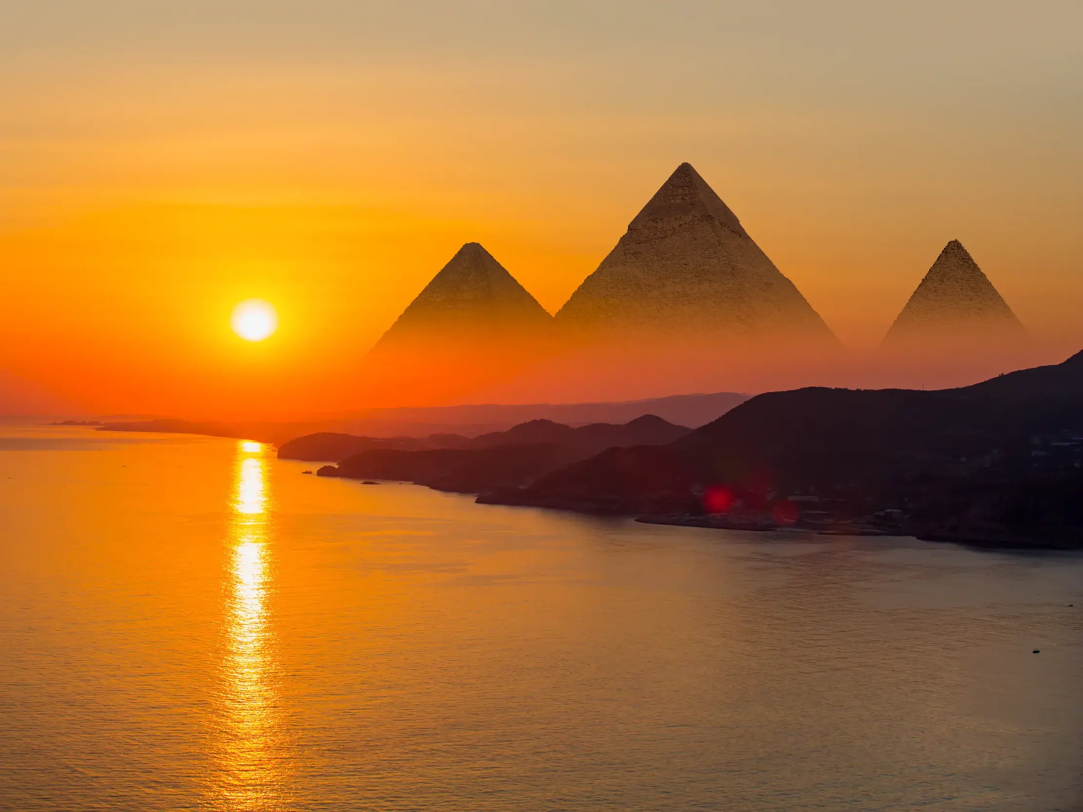Leuchtender Sonnenuntergang über dem Nil mit Blick auf die majestätischen Silhouetten der Pyramiden von Gizeh am Horizont.