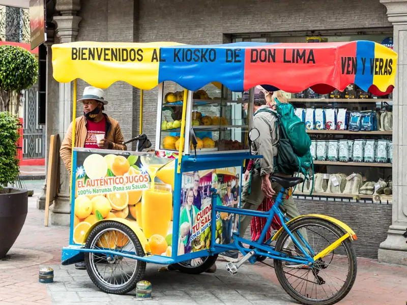 Ein Reisender mit Rucksack bestellt frischen Saft an einem farbenfrohen, mobilen Verkaufsstand in der Altstadt von Quito.