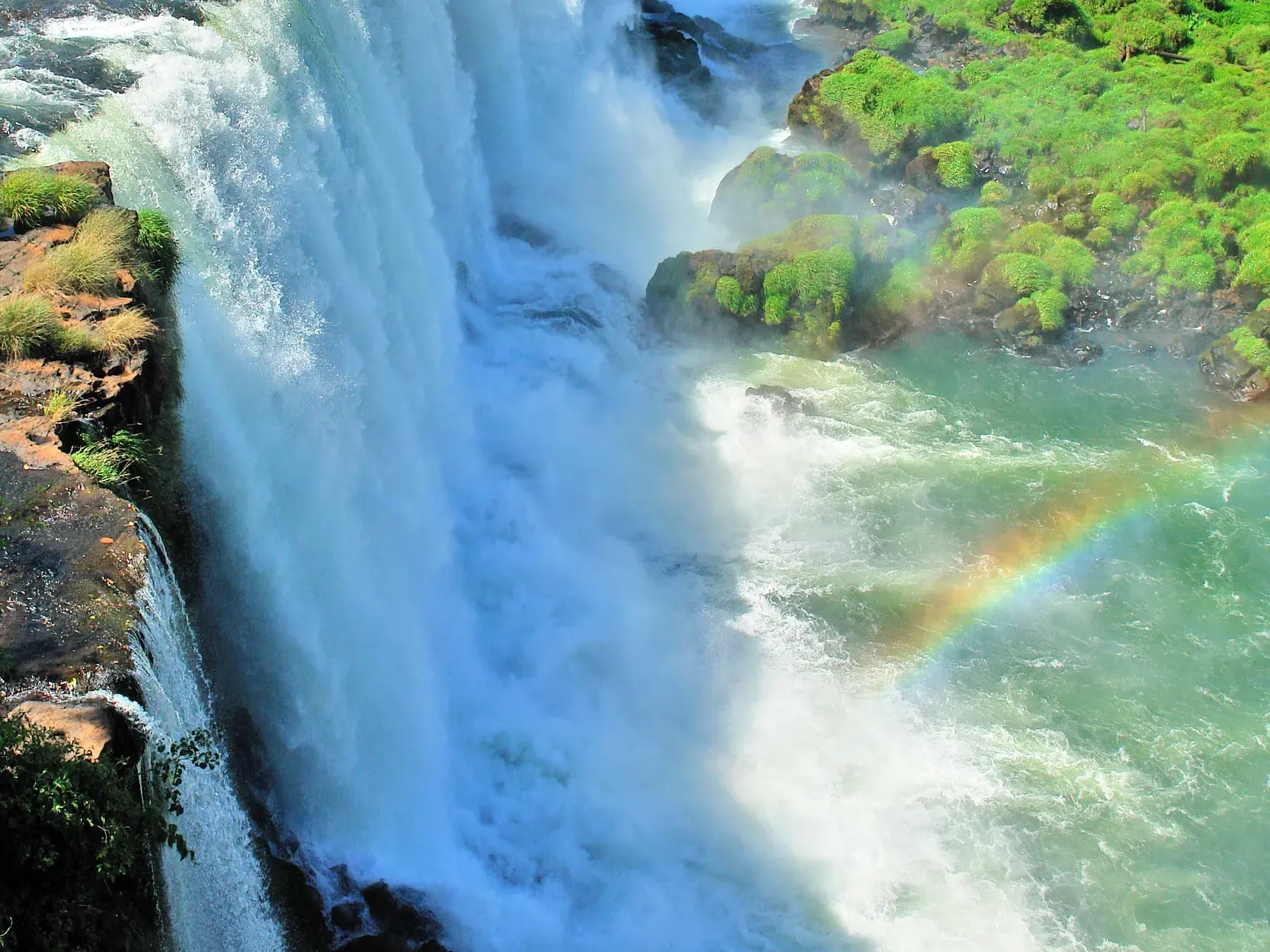 Gewaltige Wassermassen der Iguazú-Wasserfälle stürzen über begrünte Felsklippen in die Tiefe, ein Regenbogen in der Gischt.