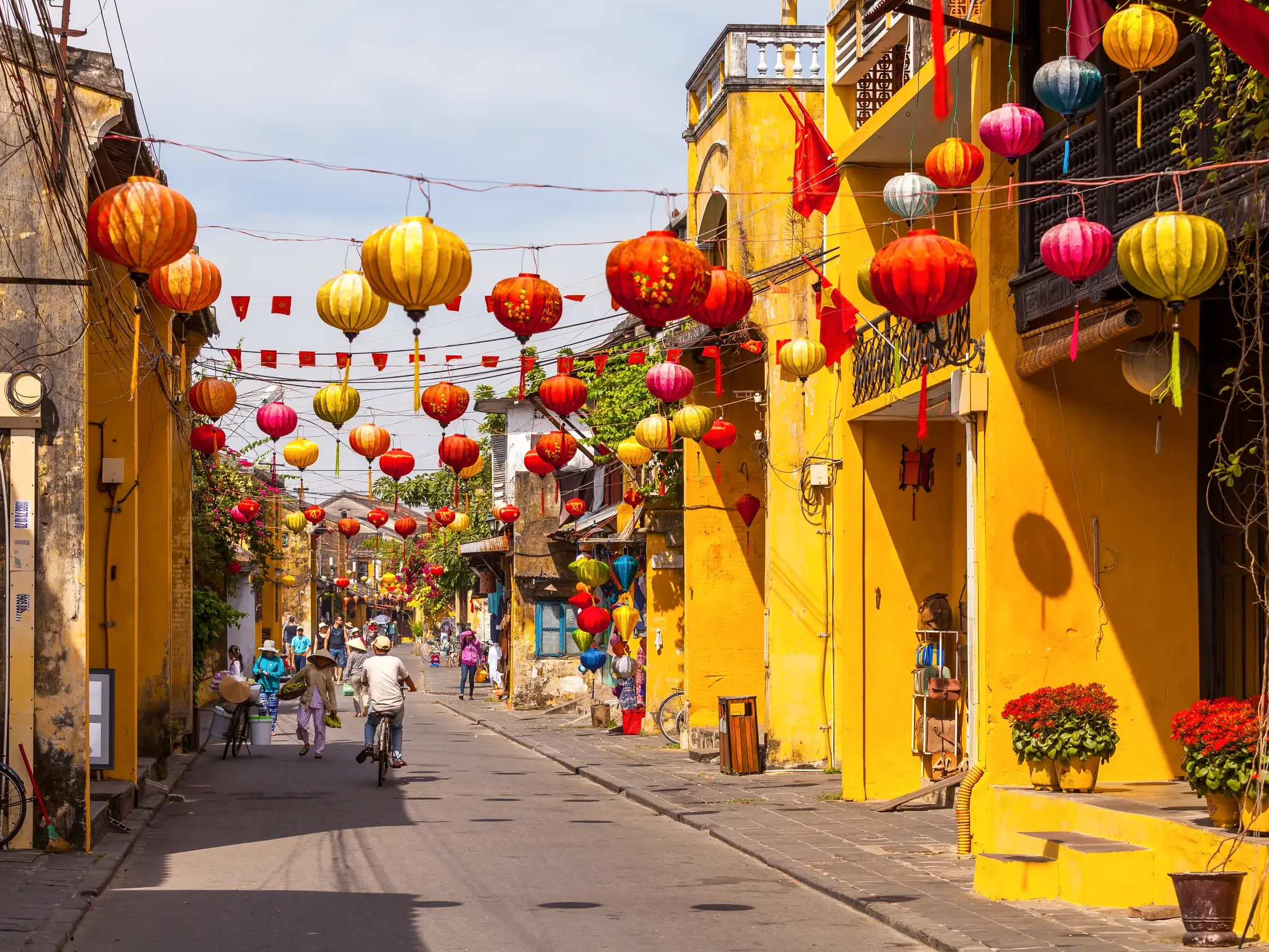 Sonnige Gasse in der Altstadt von Hoi An, geschmückt mit bunten Lampions über den Köpfen von Passanten und Radfahrern.