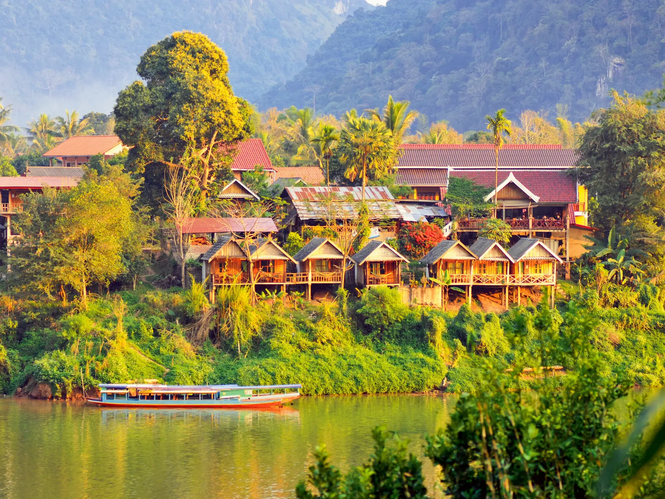 Malerisches Dorf Muang Ngoy am Ufer des Nam Ou Flusses in Laos mit Holzhütten und einem Langboot vor dichter Dschungelkulisse.