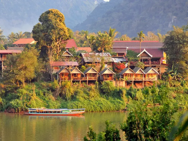 Malerisches Dorf Muang Ngoy am Ufer des Nam Ou Flusses in Laos mit Holzhütten und einem Langboot vor dichter Dschungelkulisse.