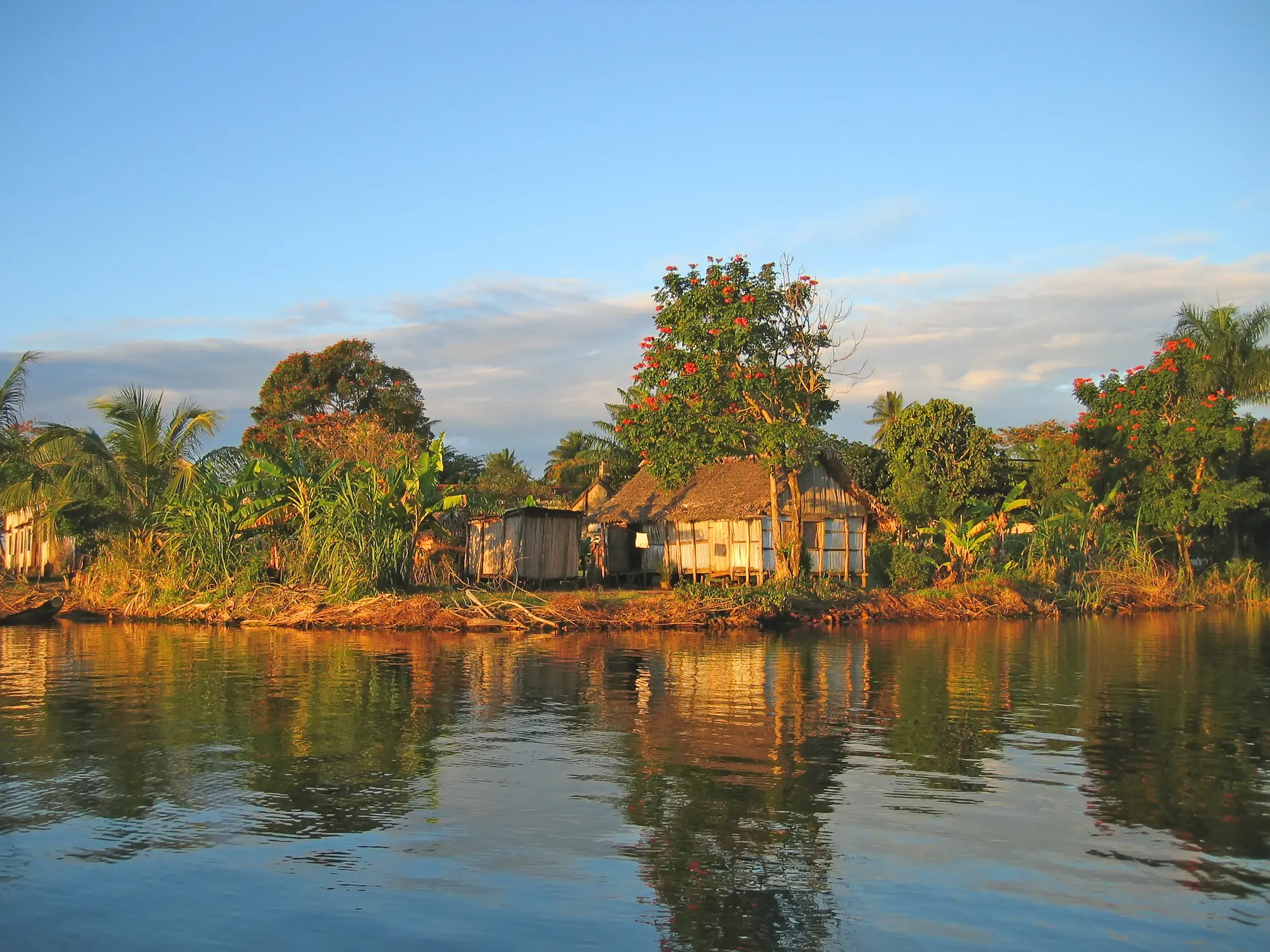 Abendsonne beleuchtet traditionelle Hütten am Ufer des Canal des Pangalanes in Madagaskar, die sich im Wasser spiegeln.