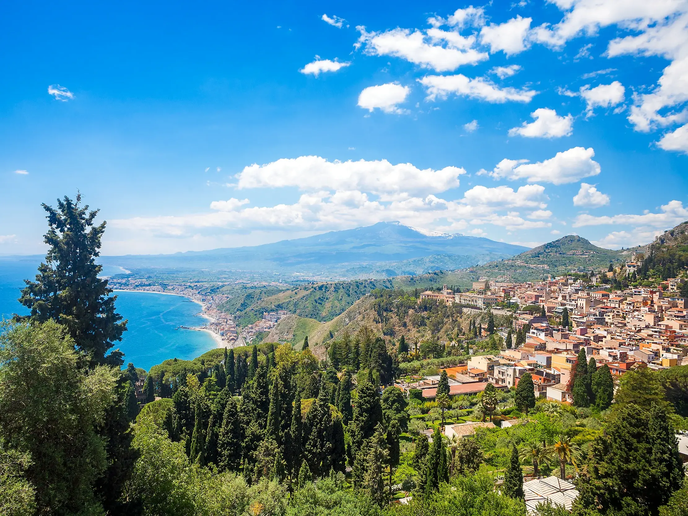 Sonniger Panoramablick auf die Stadt Taormina an der Küste Siziliens und den imposanten Vulkan Ätna unter blauem Himmel.
