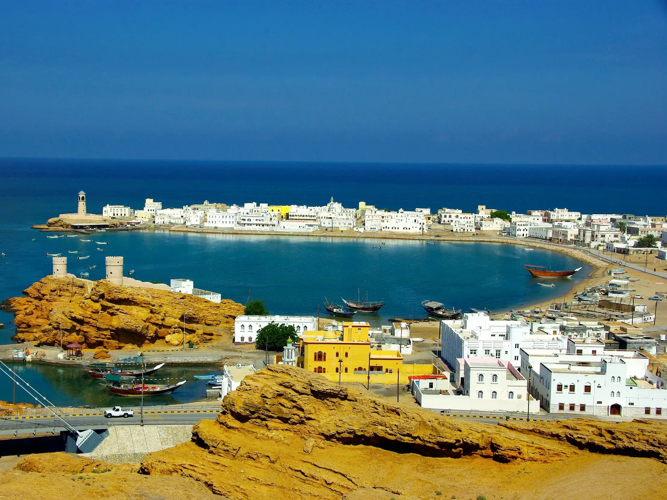 Panoramablick auf die weiße Küstenstadt Sur im Oman mit türkisblauer Bucht, historischen Wachtürmen und Dhaus.