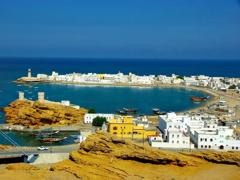 Panoramablick auf die weiße Küstenstadt Sur im Oman mit türkisblauer Bucht, historischen Wachtürmen und Dhaus.