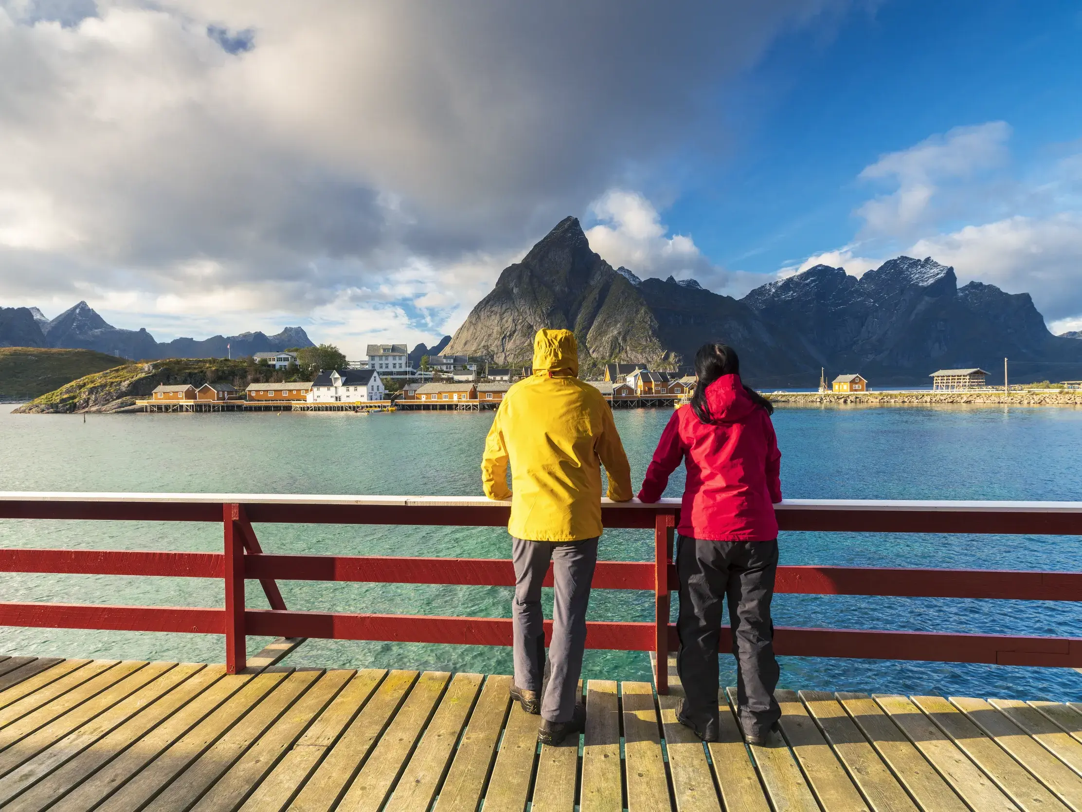 Zwei Reisende einer Studienreise genießen von einer Brücke den Blick auf das Fischerdorf Sakrisøy vor der Bergkulisse der Lofoten.