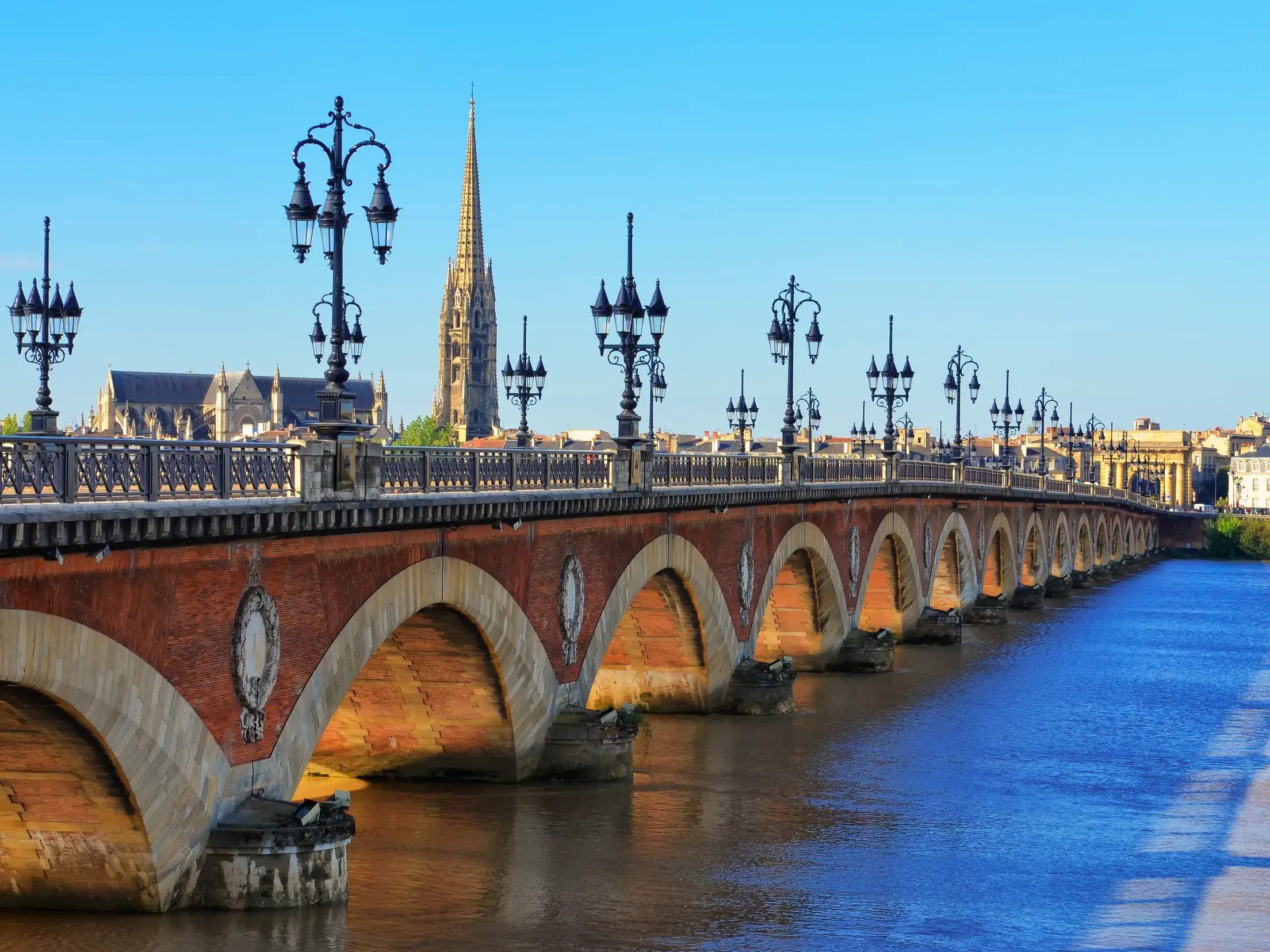 Die historische Bogenbrücke über die Garonne in Bordeaux, gesäumt von verzierten Laternen vor der Skyline mit einem Kirchturm.