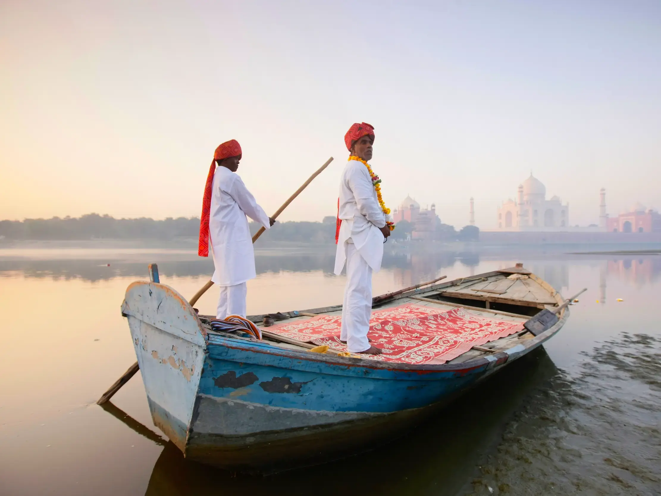 Zwei indische Männer in traditioneller Kleidung auf einem Holzboot auf dem Yamuna Fluss bei Agra, im Hintergrund das Taj Mahal.