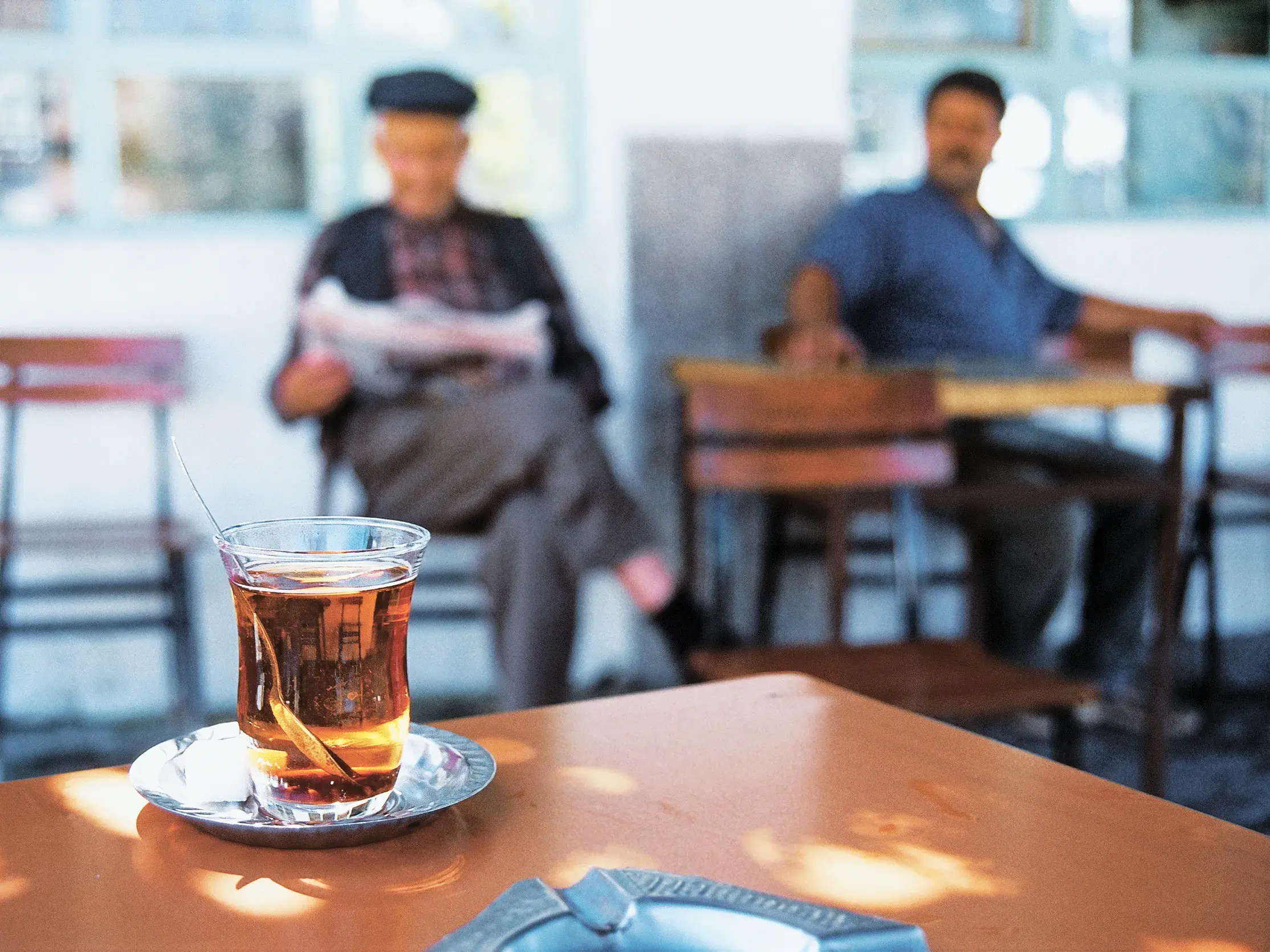 Ein Glas türkischer Tee auf einem Tisch im Teehaus, im Hintergrund entspannen zwei Männer bei ihrer täglichen Zeitungspause.