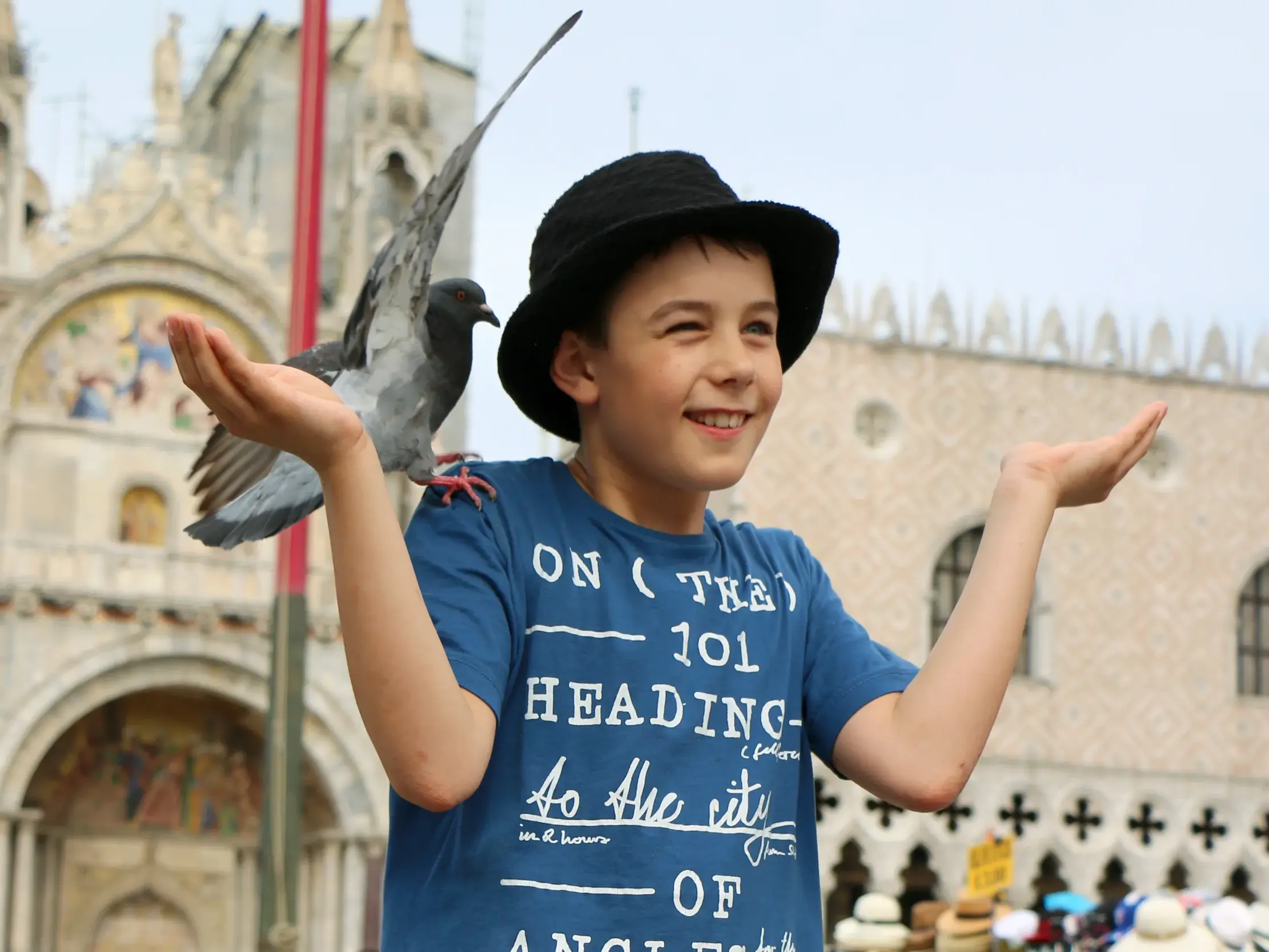 Ein lachender Junge mit Hut hält eine Taube auf seiner Hand auf dem Markusplatz in Venedig vor der Markuskirche.