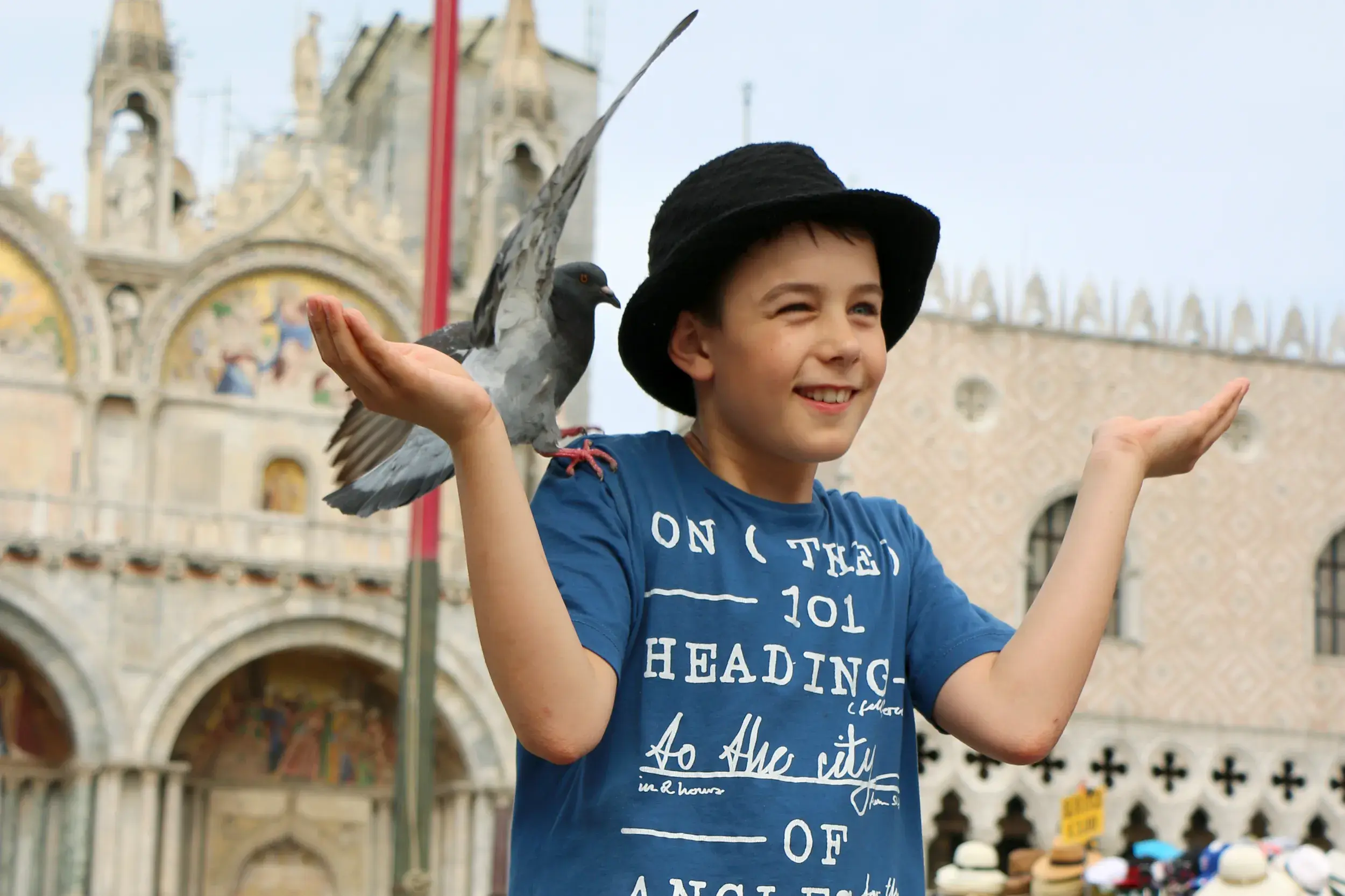 Ein lachender Junge mit Hut hält eine Taube auf seiner Hand auf dem Markusplatz in Venedig vor der Markuskirche.