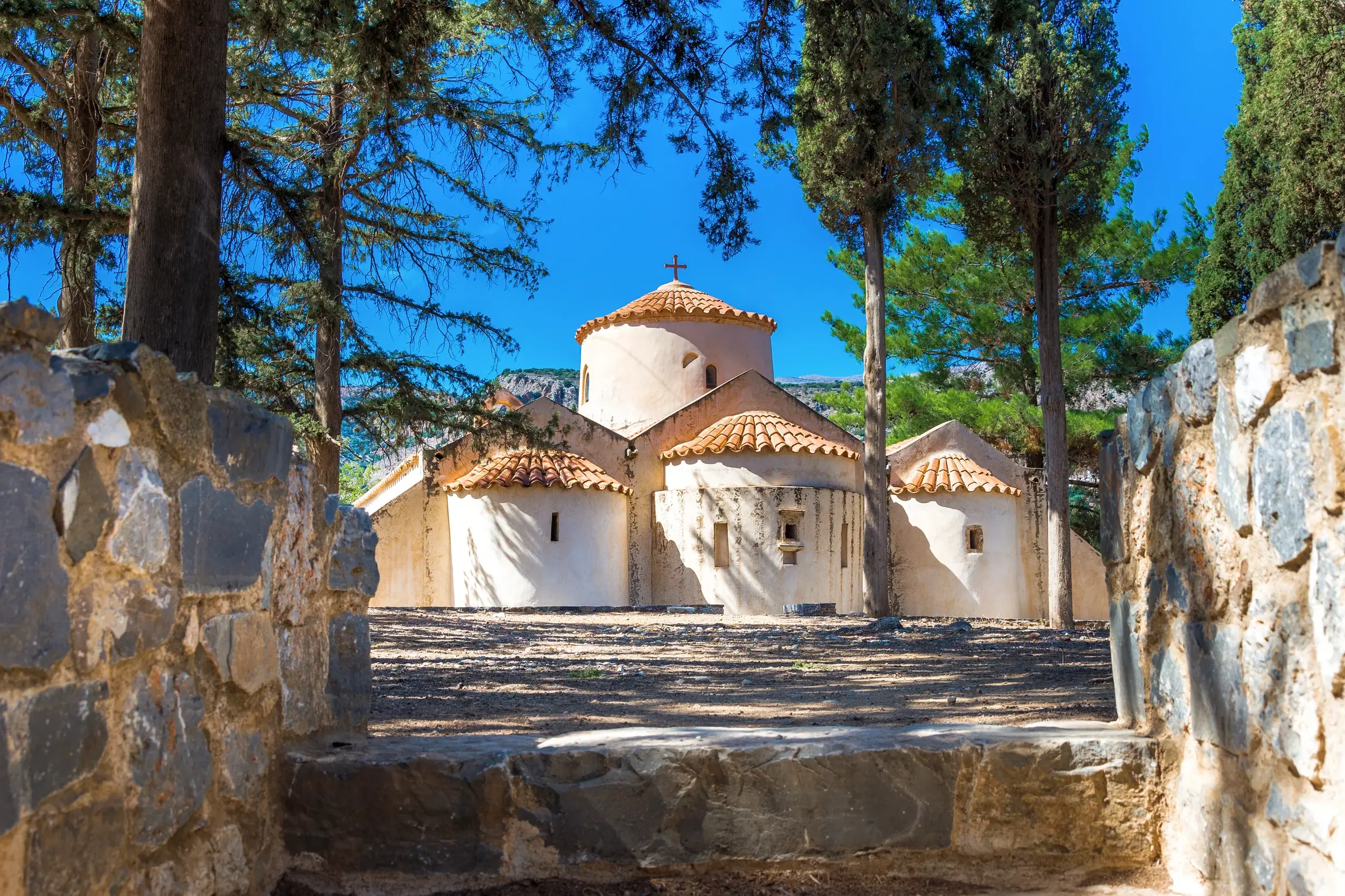 Die byzantinische Kirche Panagia Kera in Kritsa auf Kreta, eingerahmt von einer Steinmauer und hohen Bäumen an einem Sonnentag.