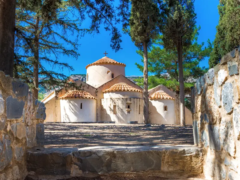 Die byzantinische Kirche Panagia Kera in Kritsa auf Kreta, eingerahmt von einer Steinmauer und hohen Bäumen an einem Sonnentag.