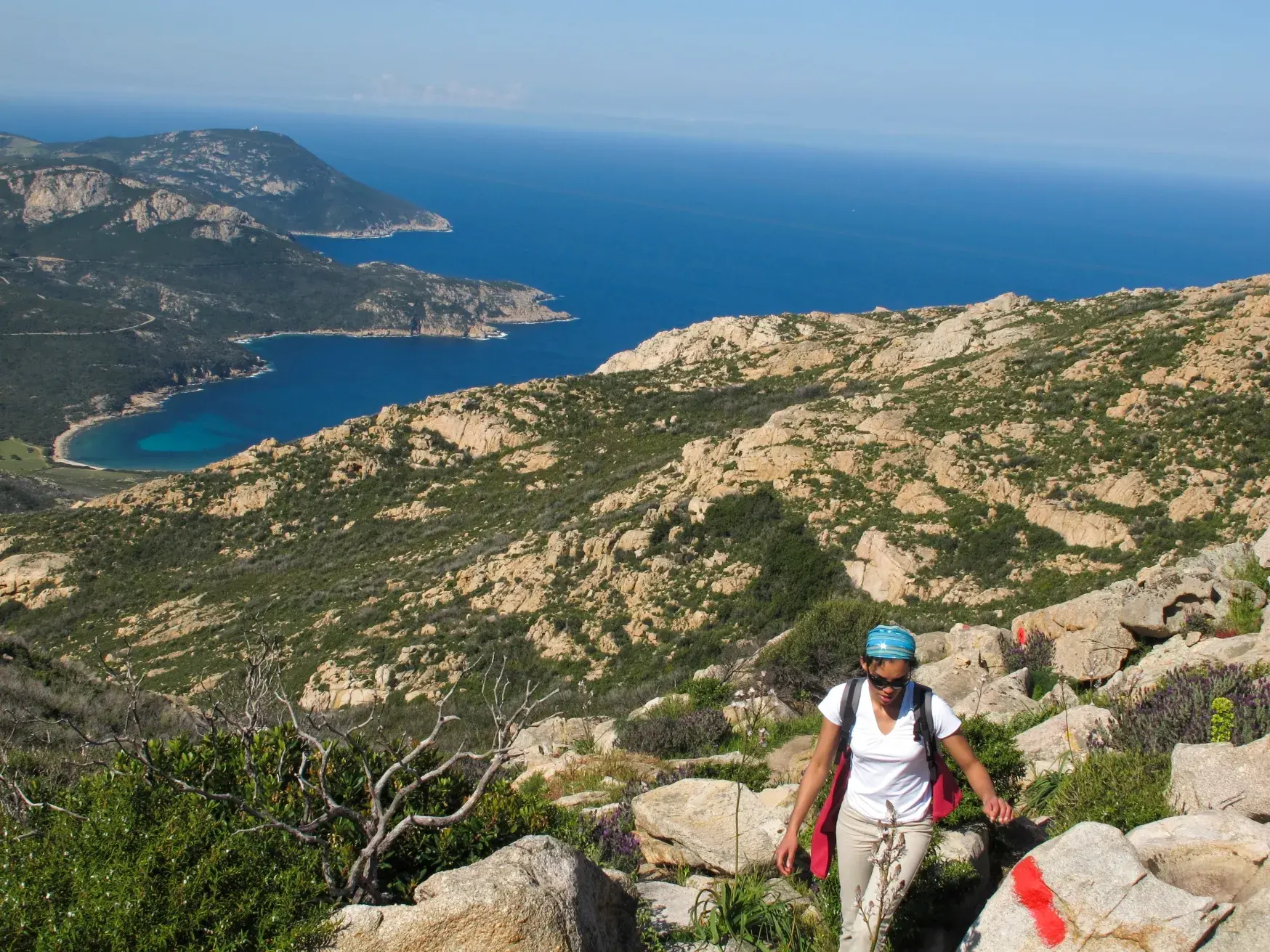 Eine Frau wandert auf einem markierten Felsenpfad durch die Macchia Korsikas mit Panoramablick über die Küste und das Meer.