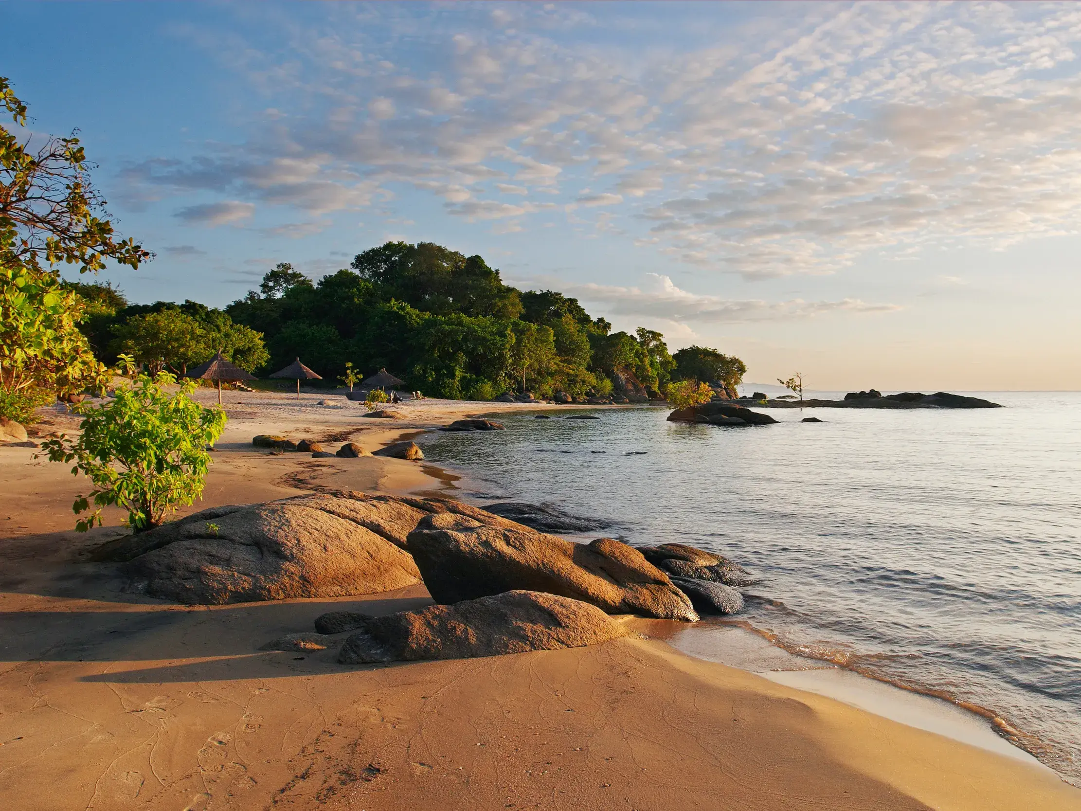 Idyllische, von Felsen und grünen Hügeln gesäumte Sandbucht am Malawi-See in der warmen Abendsonne Malawis.