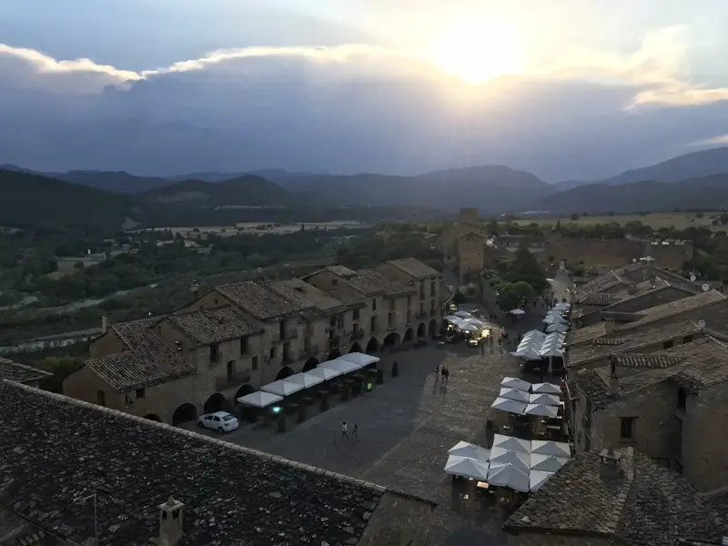 Blick über die Dächer des mittelalterlichen Dorfes Alquézar auf den Dorfplatz bei Sonnenuntergang mit Bergkulisse der Pyrenäen.