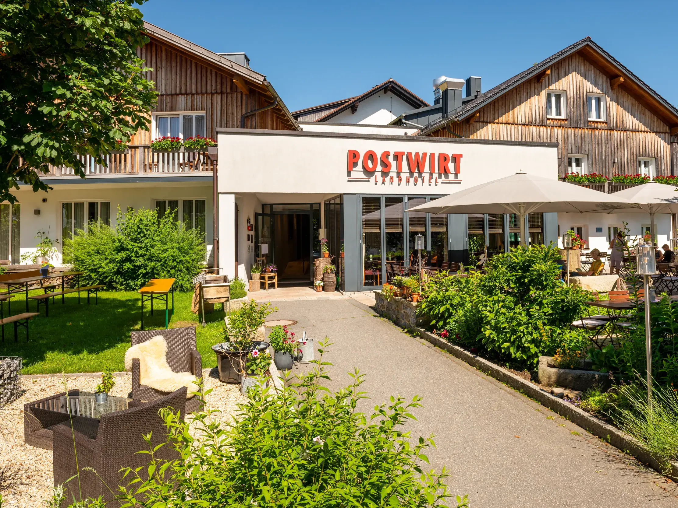 Sonnige Außenansicht des Postwirt Landhotel mit gemütlicher Terrasse, grüner Gartenanlage und traditioneller Holzfassade.