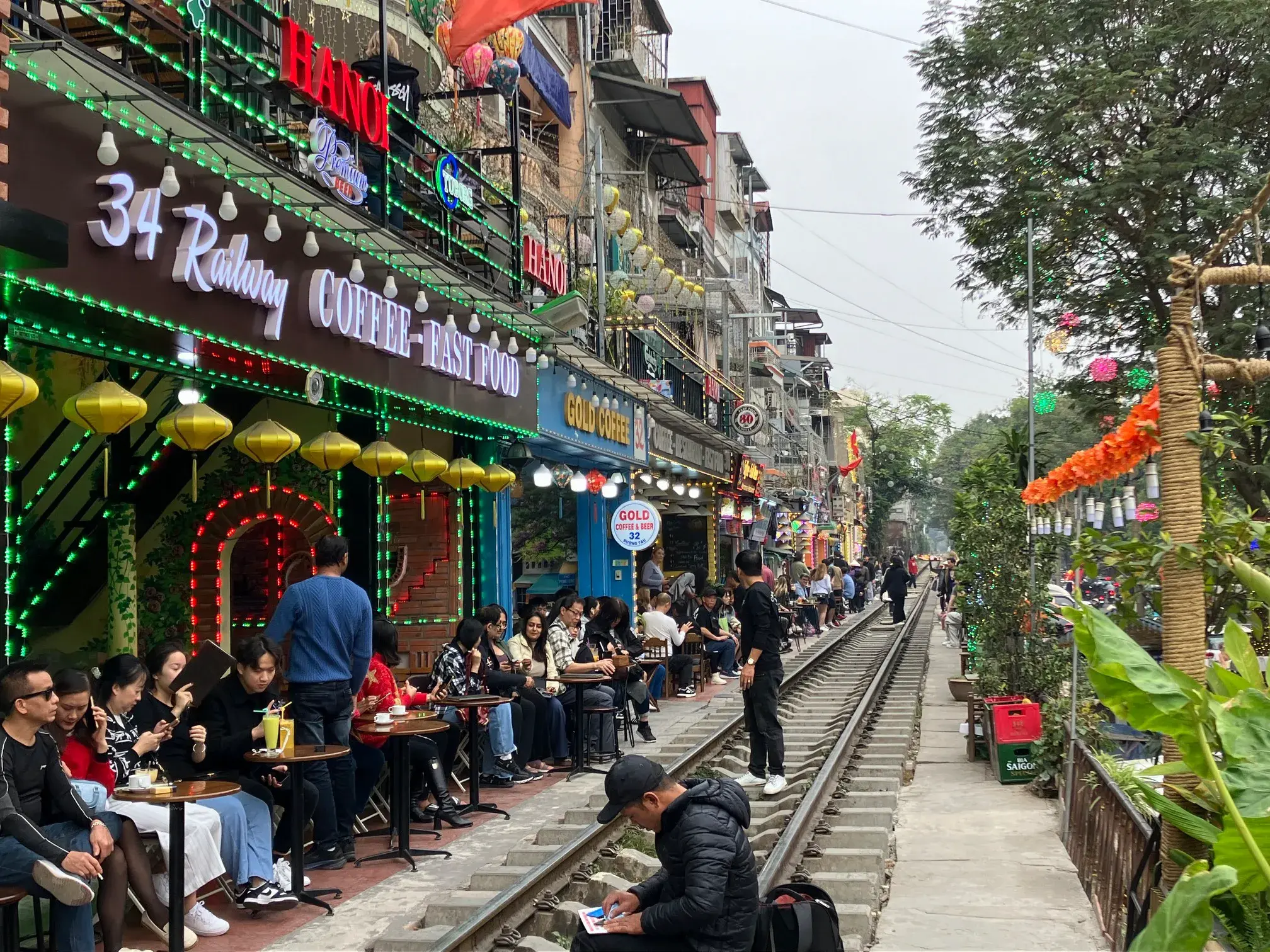 Menschen sitzen in belebten Cafés mit gelben Lampions direkt an den Gleisen der berühmten Train Street in Hanoi.