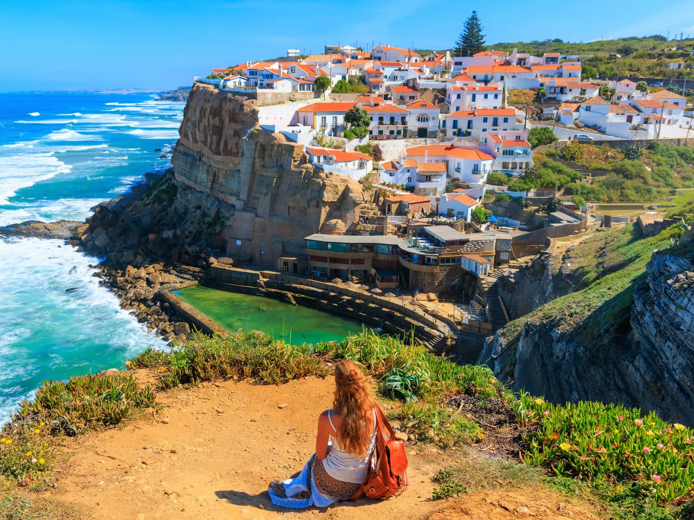 Eine Reisende blickt von einer Klippe auf das malerische Dorf an der Steilküste nahe Sintra und den weiten Atlantik.