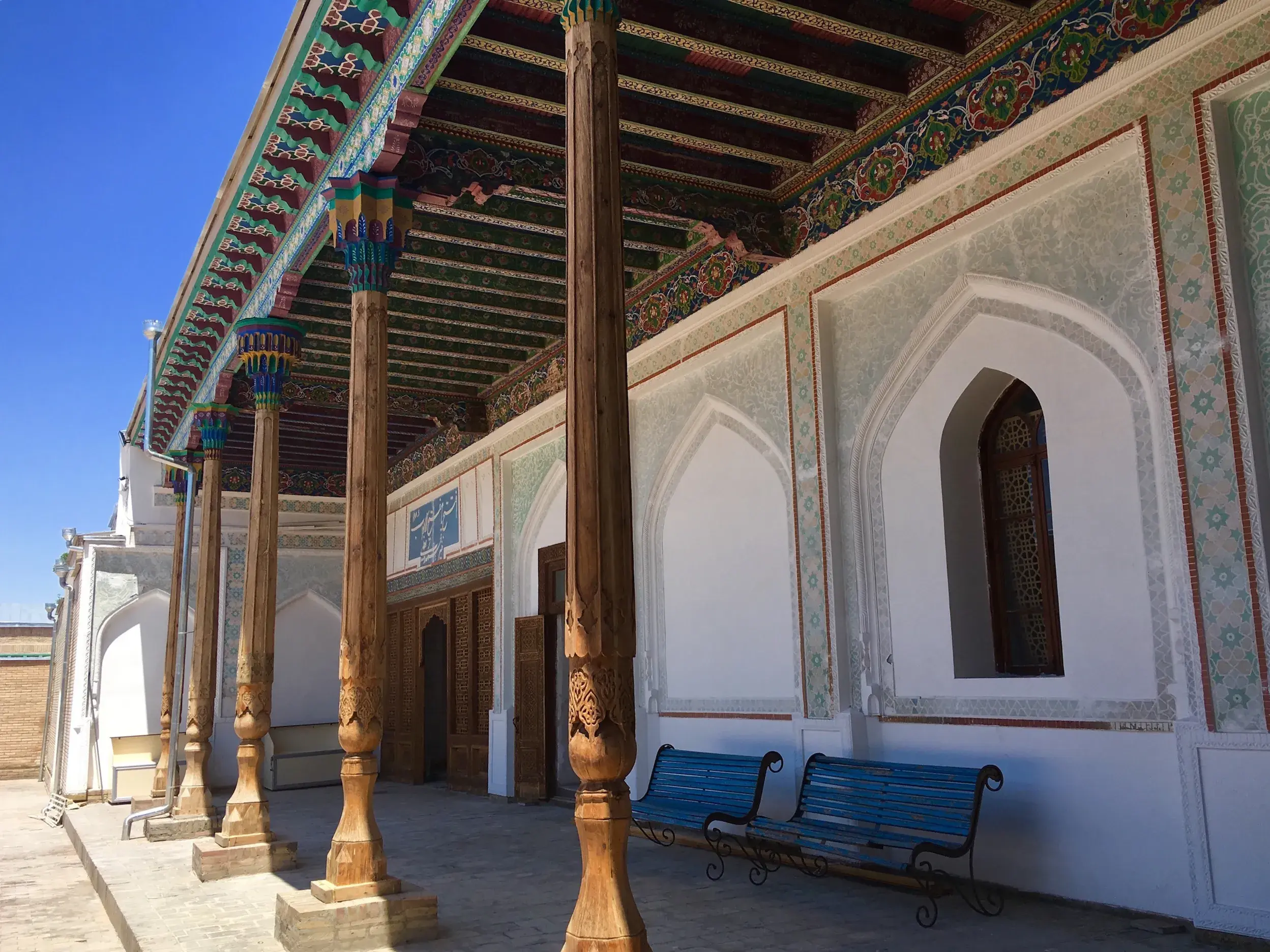 Kunstvoll bemalte Decke und geschnitzte Holzsäulen der Vorhalle der Bolo-Hovuz-Moschee in Buchara bei hellem Tageslicht.