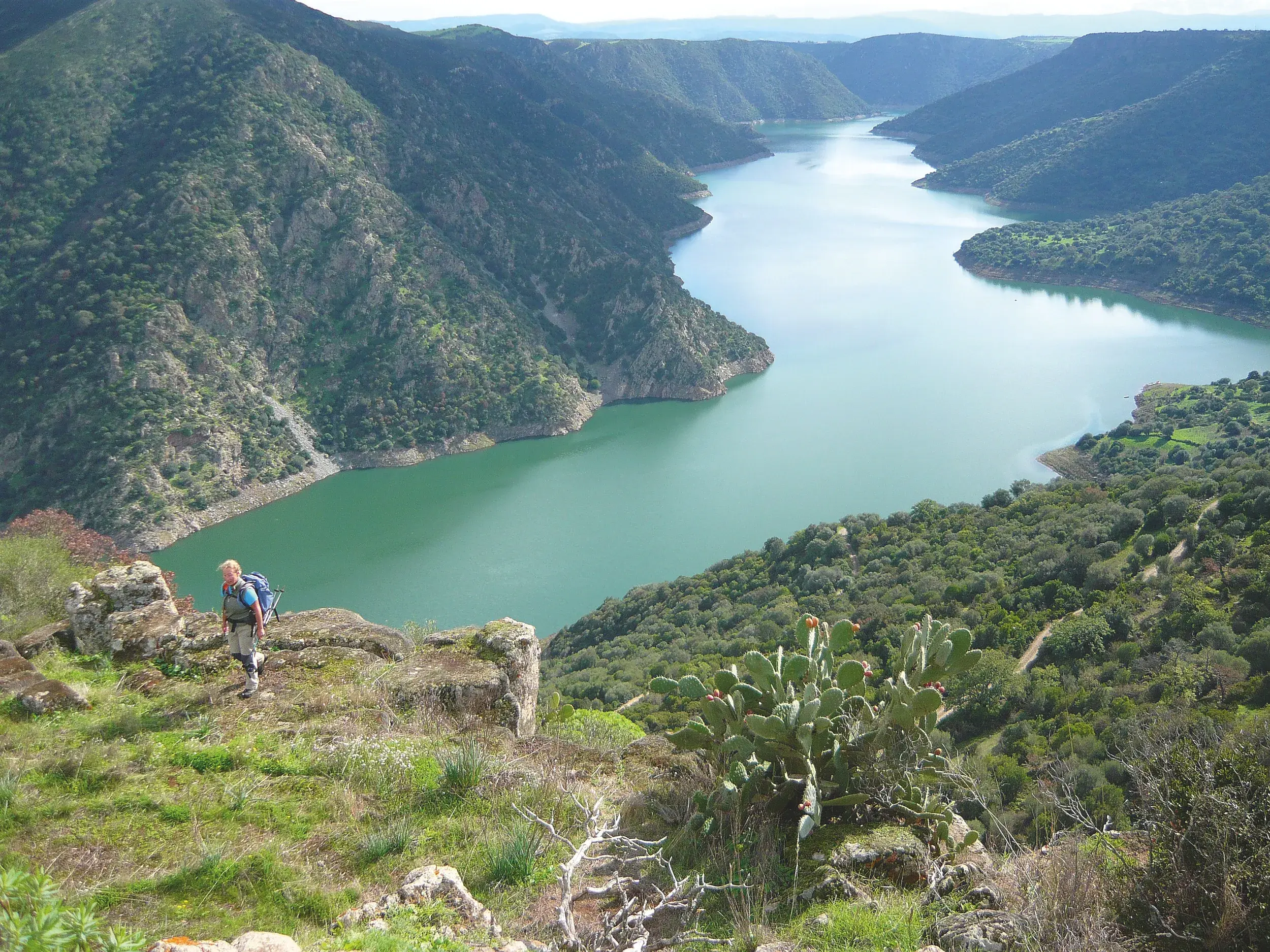 Wanderer mit Rucksack auf einem Felsvorsprung mit Blick auf einen fjordartigen, grünen Stausee in der Berglandschaft von Austis.