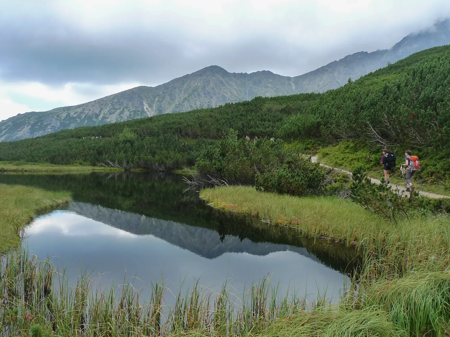 Zwei Wanderer auf einem schmalen Pfad entlang eines ruhigen Bergsees, in dem sich die umliegenden Gipfel der Hohen Tatra spiegeln.