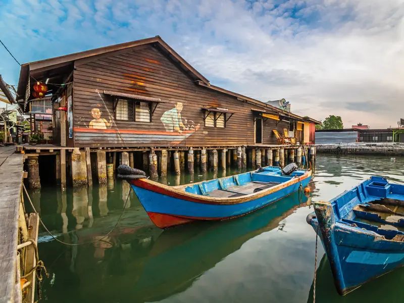 Zwei blaue Fischerboote dümpeln im Wasser vor einem hölzernen Stelzenhaus der Clan Jetties in George Town auf Penang.
