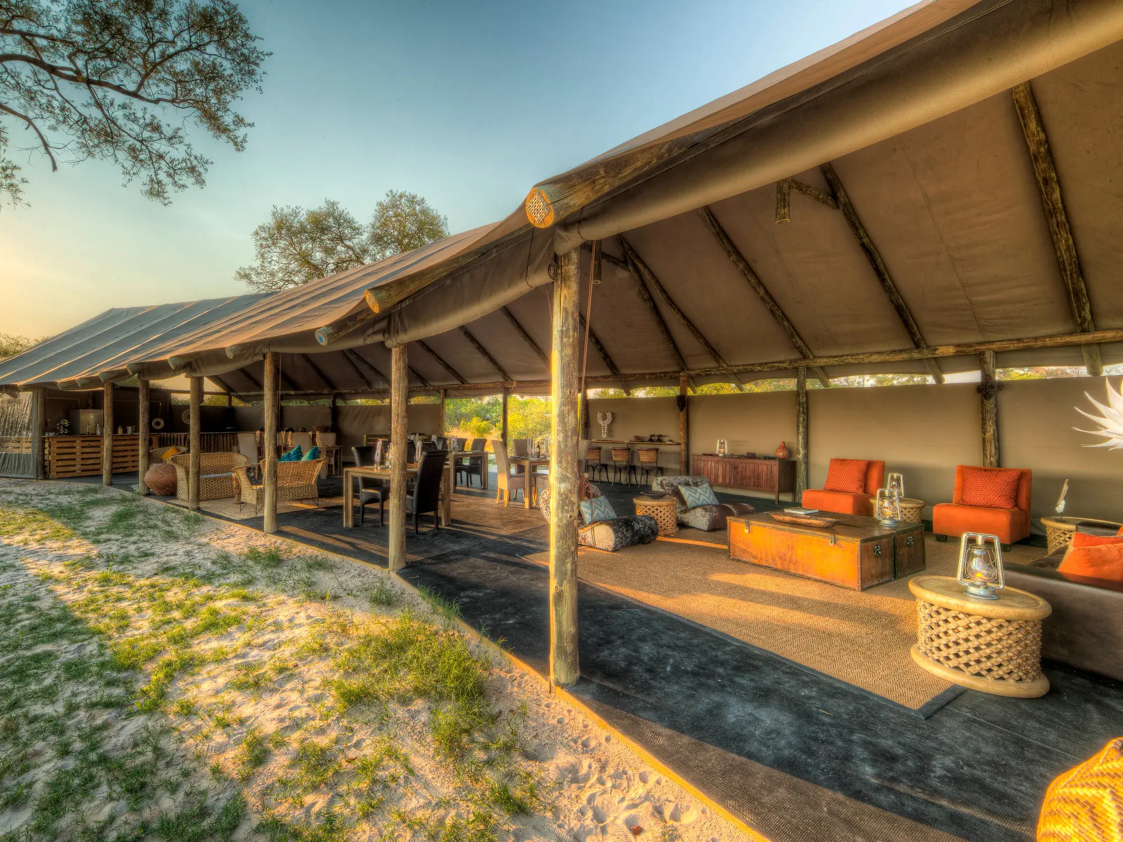 Offene Lounge des Khwai River View Camp in Botswana mit Sesseln und Essbereich auf einer Holzterrasse bei Sonnenuntergang.
