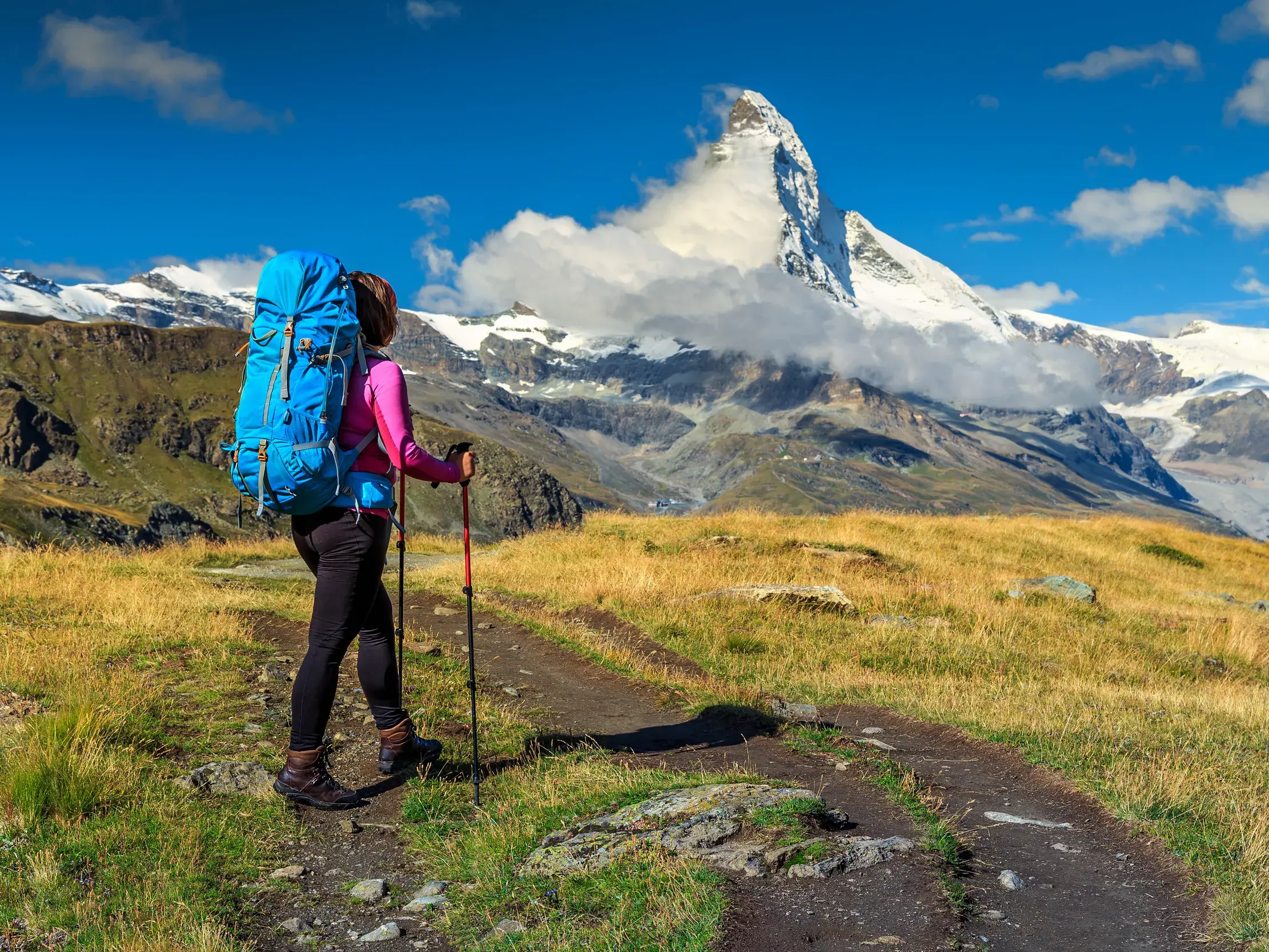Eine Wanderin mit Trekkingrucksack pausiert auf einem Höhenweg in den Walliser Bergen und blickt auf das Matterhorn.