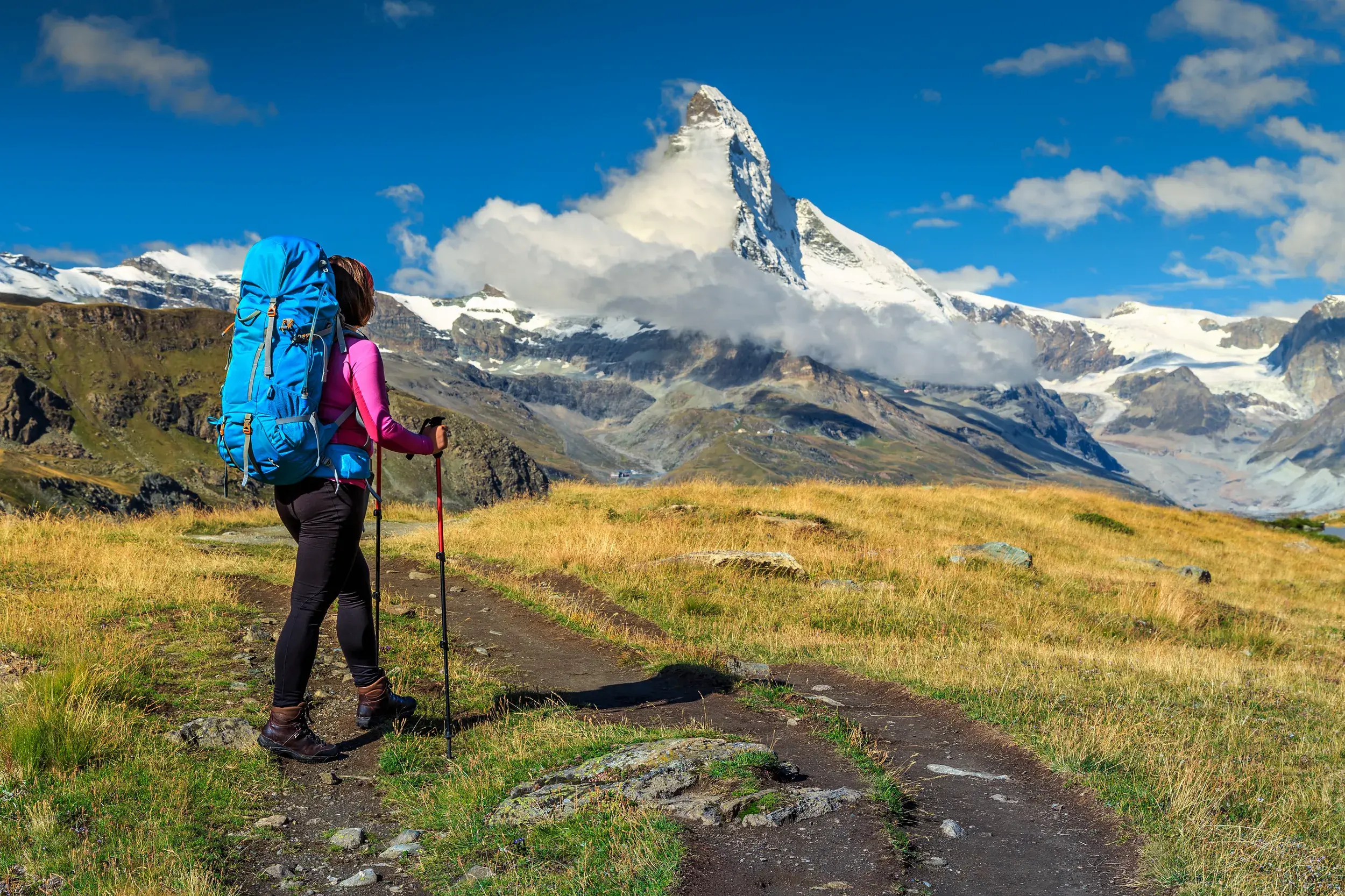 Eine Wanderin mit Trekkingrucksack pausiert auf einem Höhenweg in den Walliser Bergen und blickt auf das Matterhorn.
