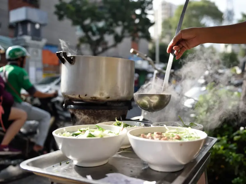Eine Hand füllt mit einer Kelle dampfende Brühe aus einem großen Topf in Schalen an einem Straßenstand in Vietnam.