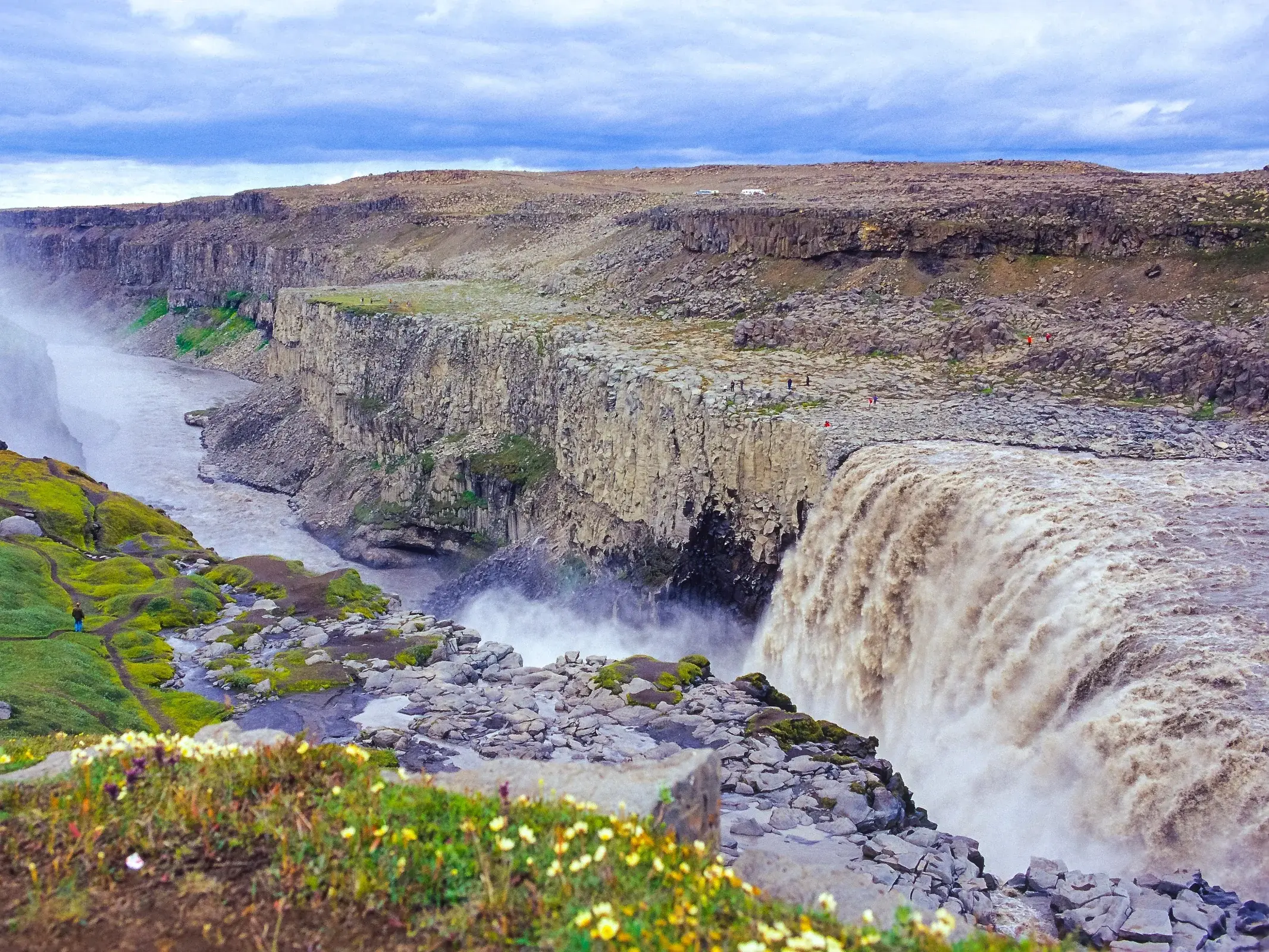 Tosende Wassermassen des Dettifoss stürzen in die Jökulsárgljúfur-Schlucht in Island, ein Wanderer am moosbewachsenen Ufer.