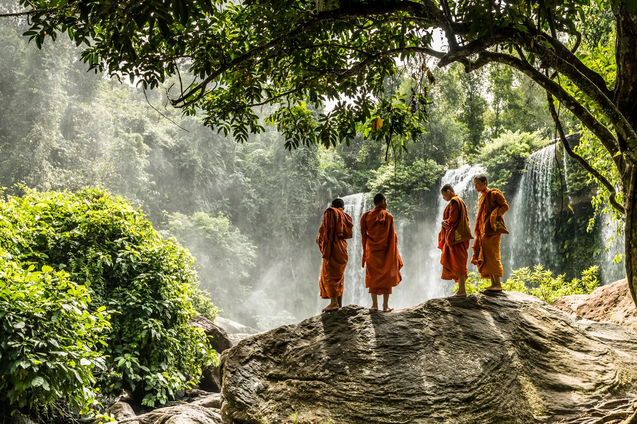 Vier buddhistische Mönche in orangen Roben stehen auf einem Felsen und betrachten den Phnom Kulen Wasserfall in Kambodscha.