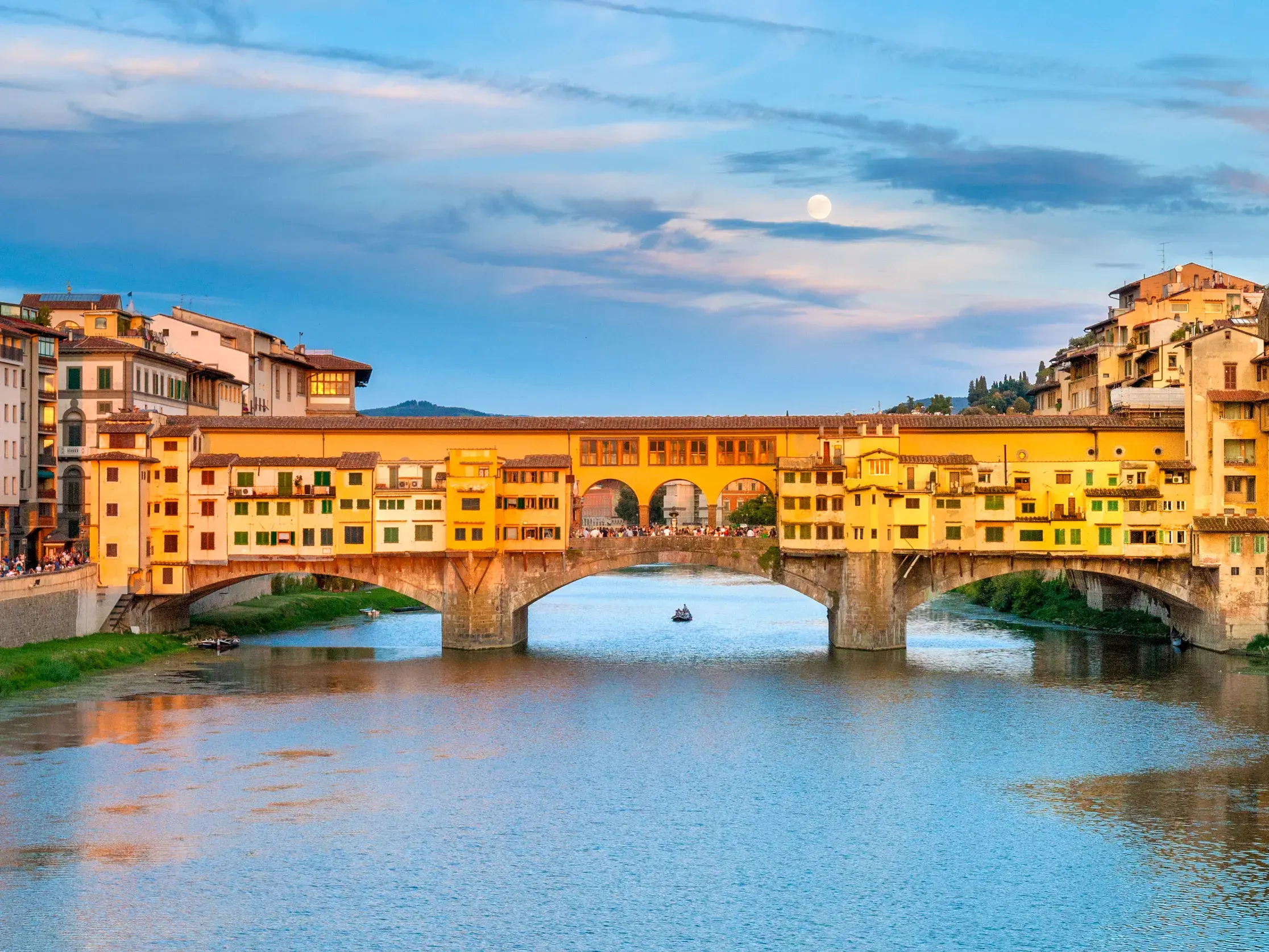 Die historische Ponte Vecchio Brücke in Florenz im goldenen Abendlicht über dem Arno, mit Vollmond am Himmel.