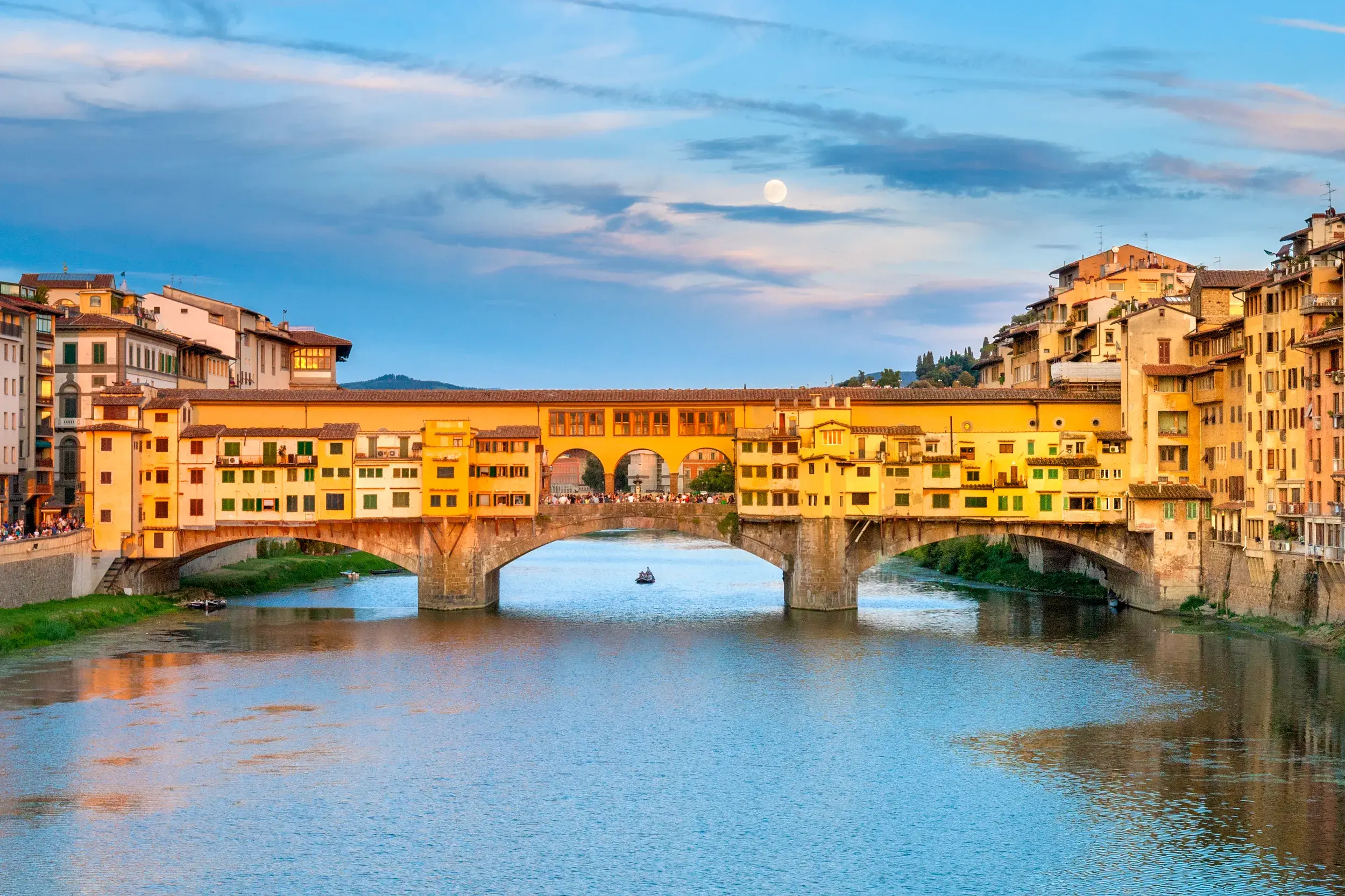 Die historische Ponte Vecchio Brücke in Florenz im goldenen Abendlicht über dem Arno, mit Vollmond am Himmel.