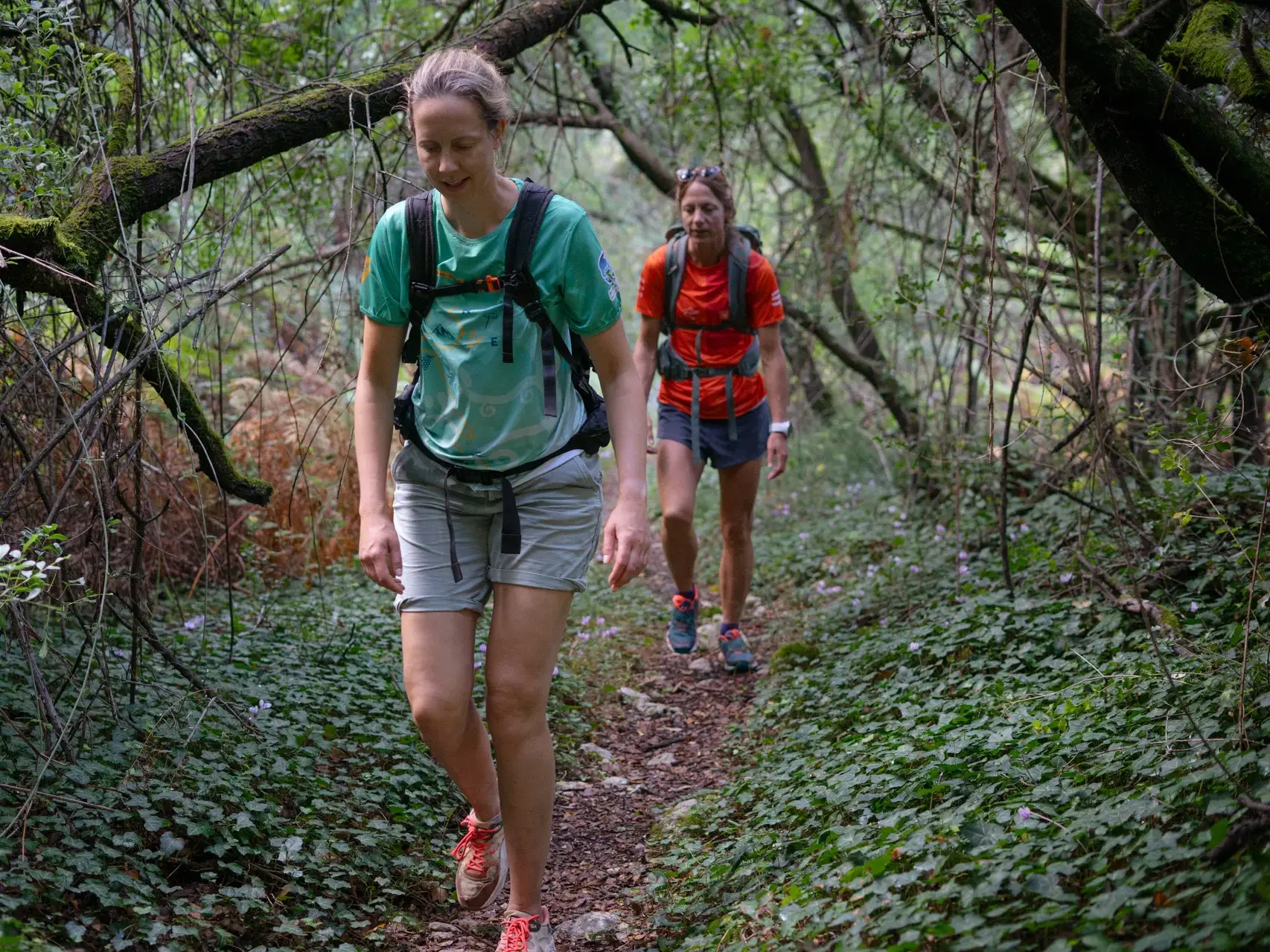 Zwei Wanderinnen mit Rucksäcken auf einem schmalen Waldpfad des Korfu-Trails, umgeben von üppiger, grüner Vegetation.